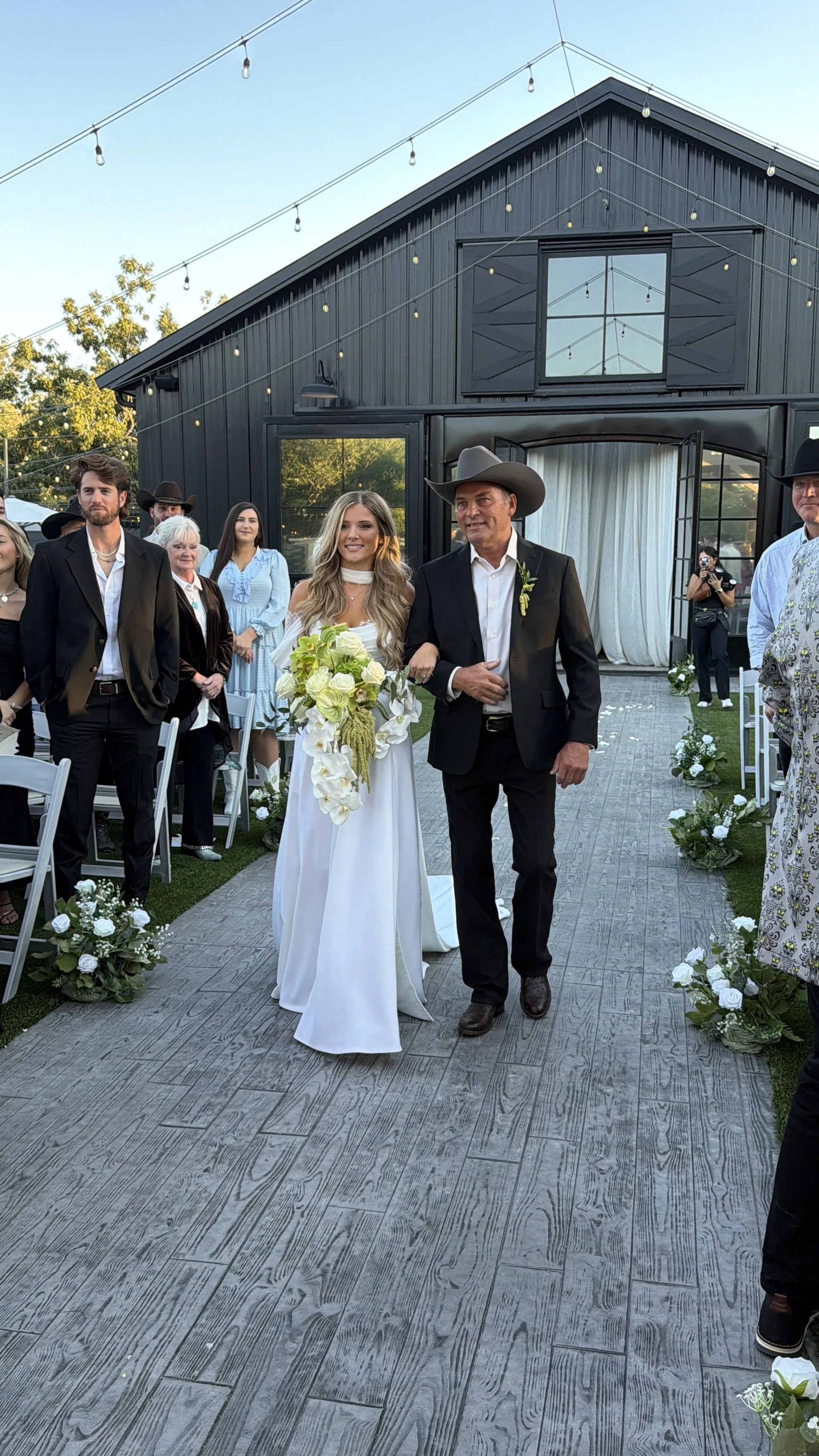 Bride walking down the aisle arm-in-arm with a man at a wedding ceremony, with guests seated and standing around, outdoor setting with string lights and a black building in the background.