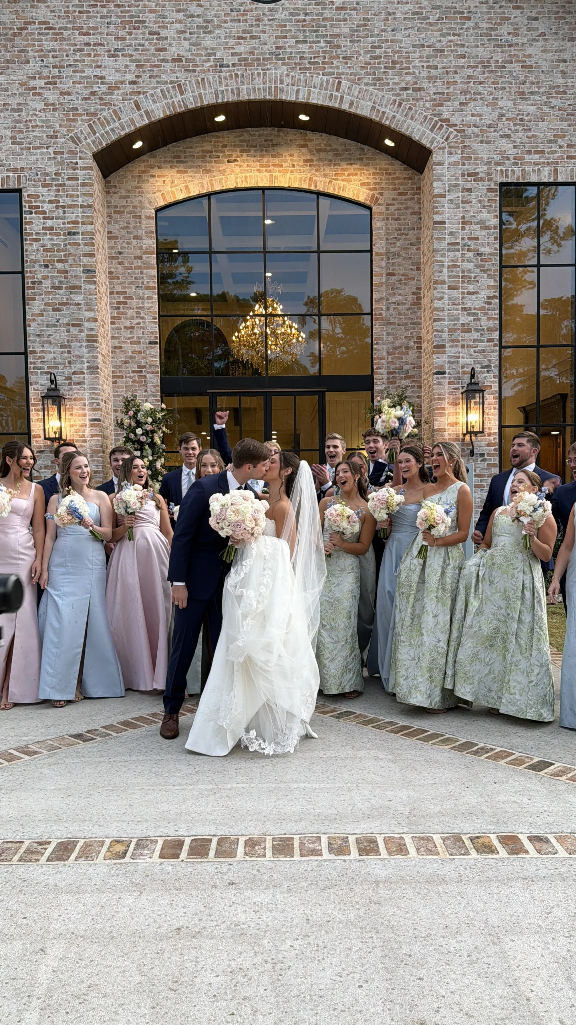 A bride and groom kissing, surrounded by wedding party members holding bouquets outside a brick building with large glass windows and a chandelier inside.