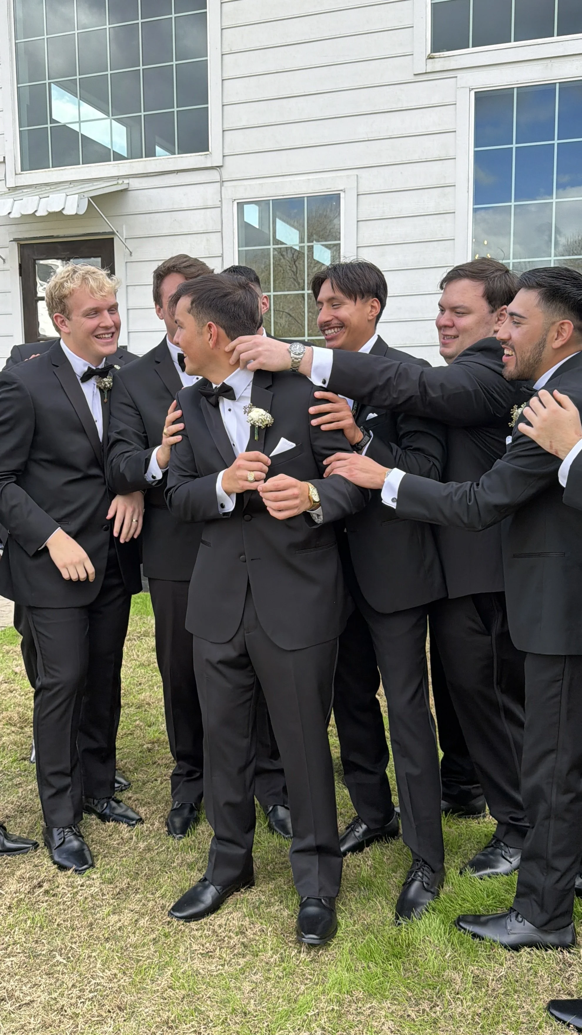 Group of men in tuxedos standing outdoors and celebrating, smiling and touching each other's shoulders.