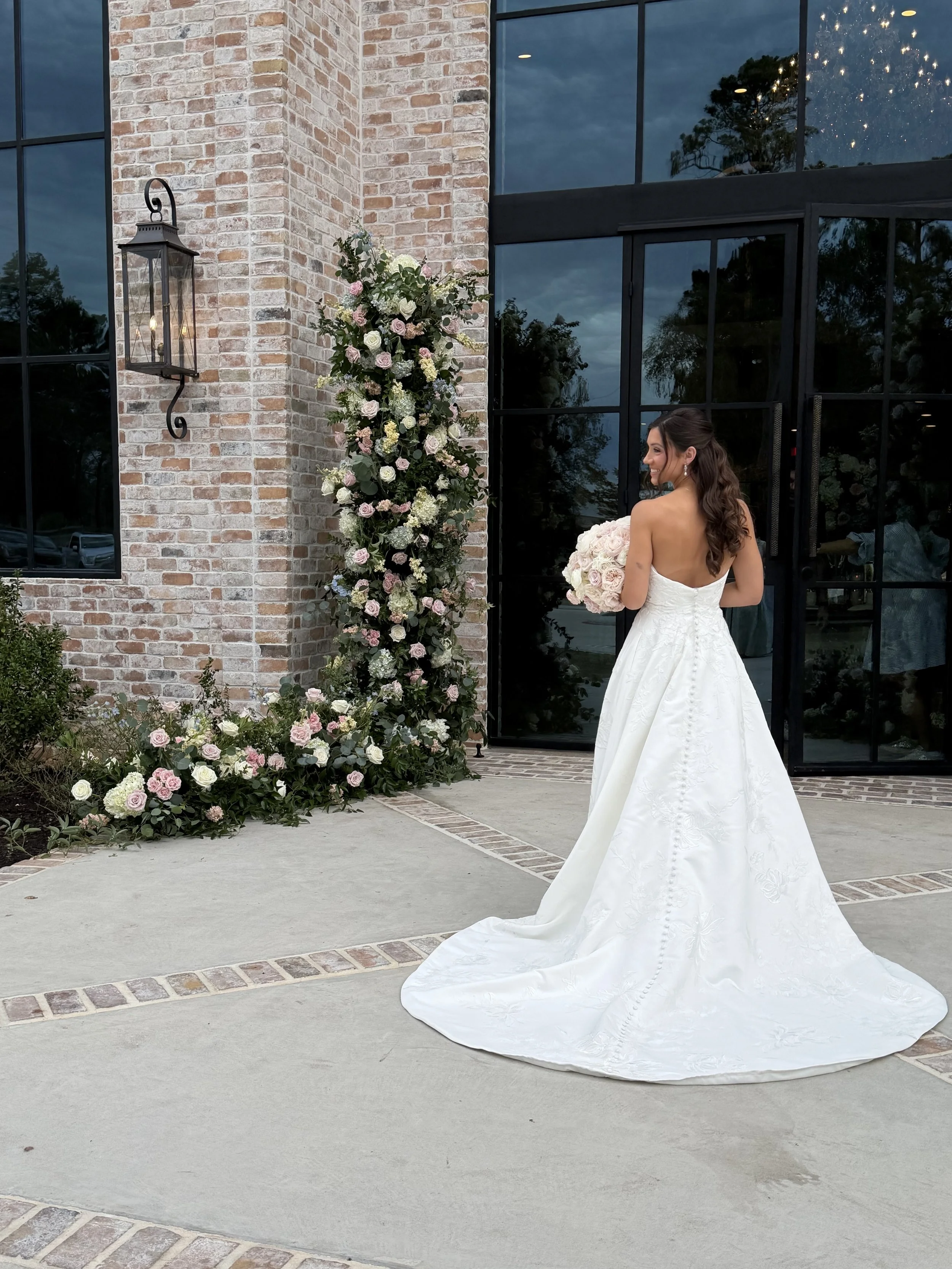 A bride in a white wedding gown with an open back, holding a bouquet of pink and white roses, standing outside a building with large glass windows and a brick wall, decorated with a floral arrangement and a black lantern.