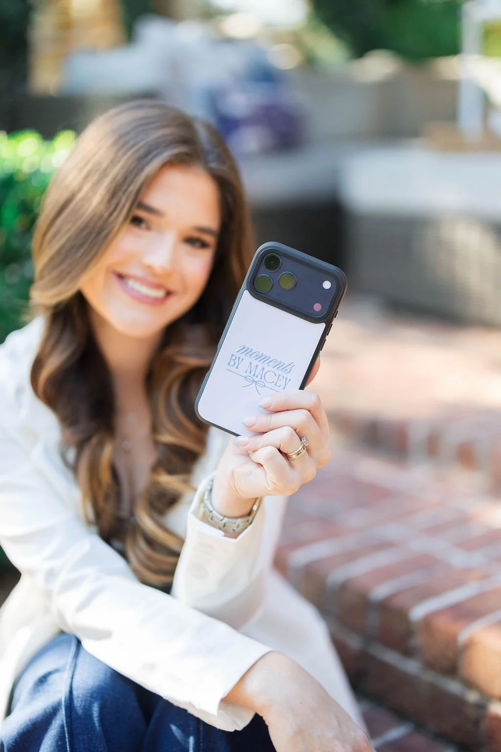 A woman with long wavy hair smiling and holding a smartphone with a case that says 'moments BY MACEY.' She is wearing a white blazer and a watch, sitting outdoors on a brick surface, with a blurred background of greenery and outdoor furniture.