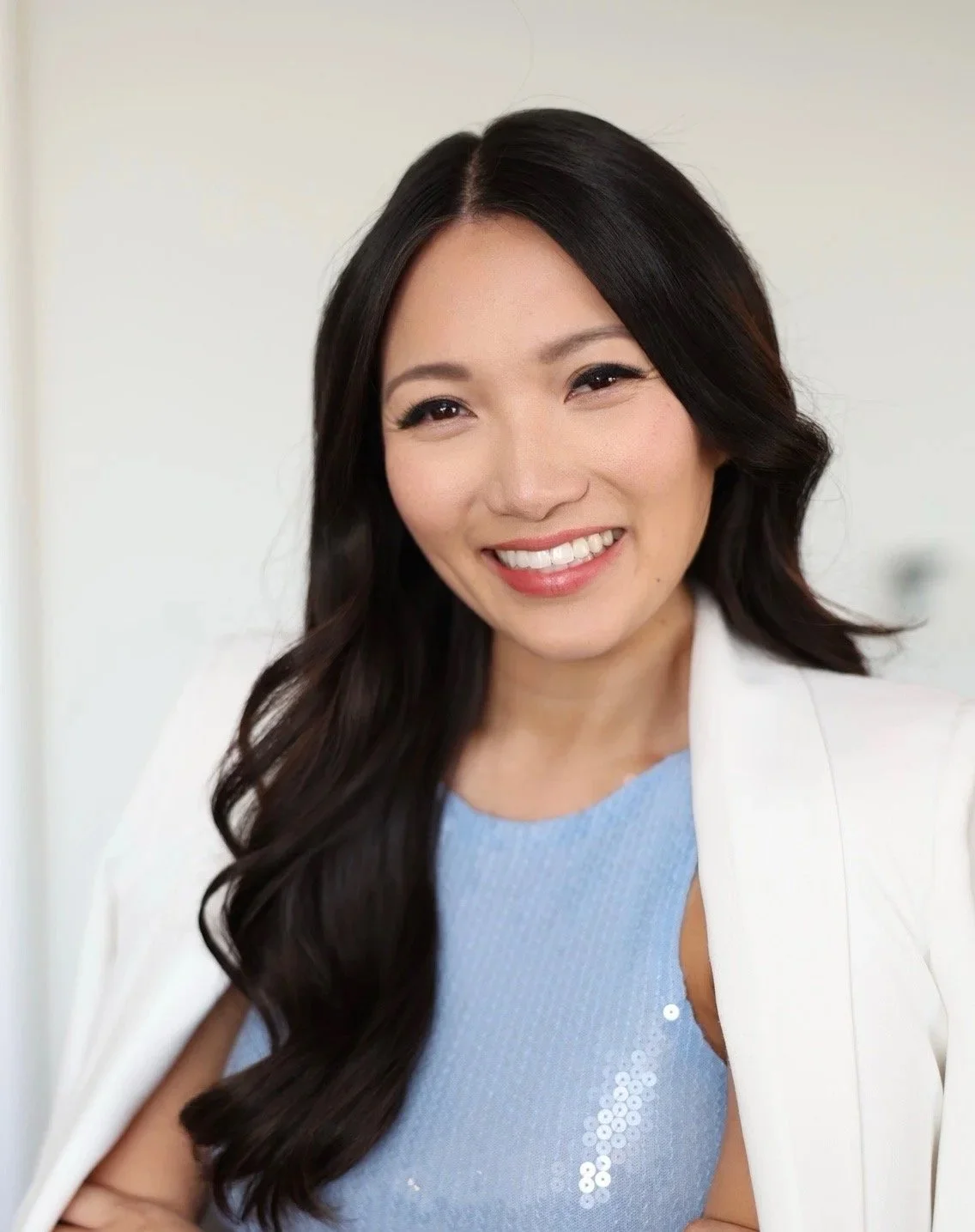 A smiling woman with long black hair wearing a light blue top and white blazer.