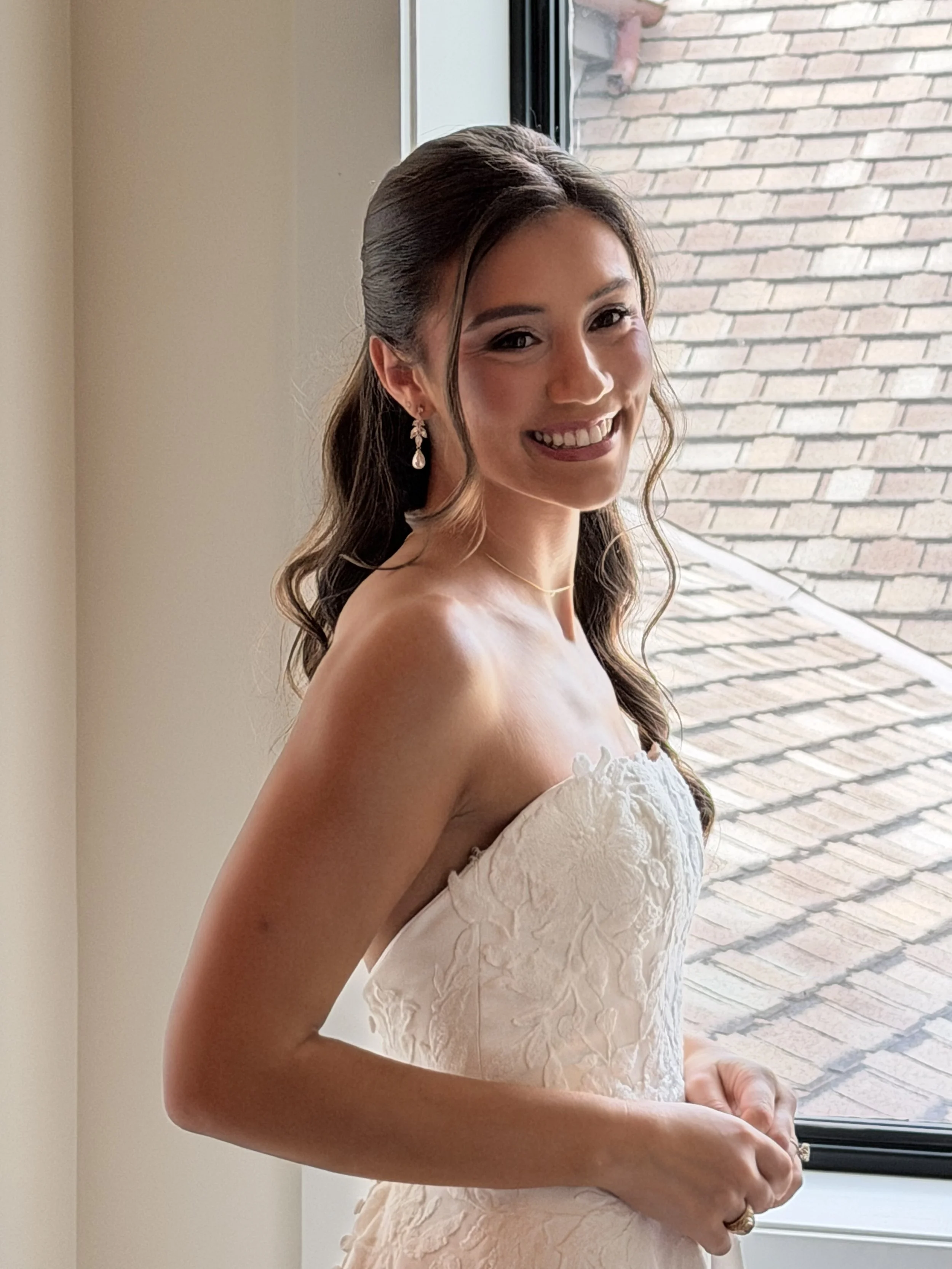 A young woman in a white strapless wedding dress standing near a window, smiling.