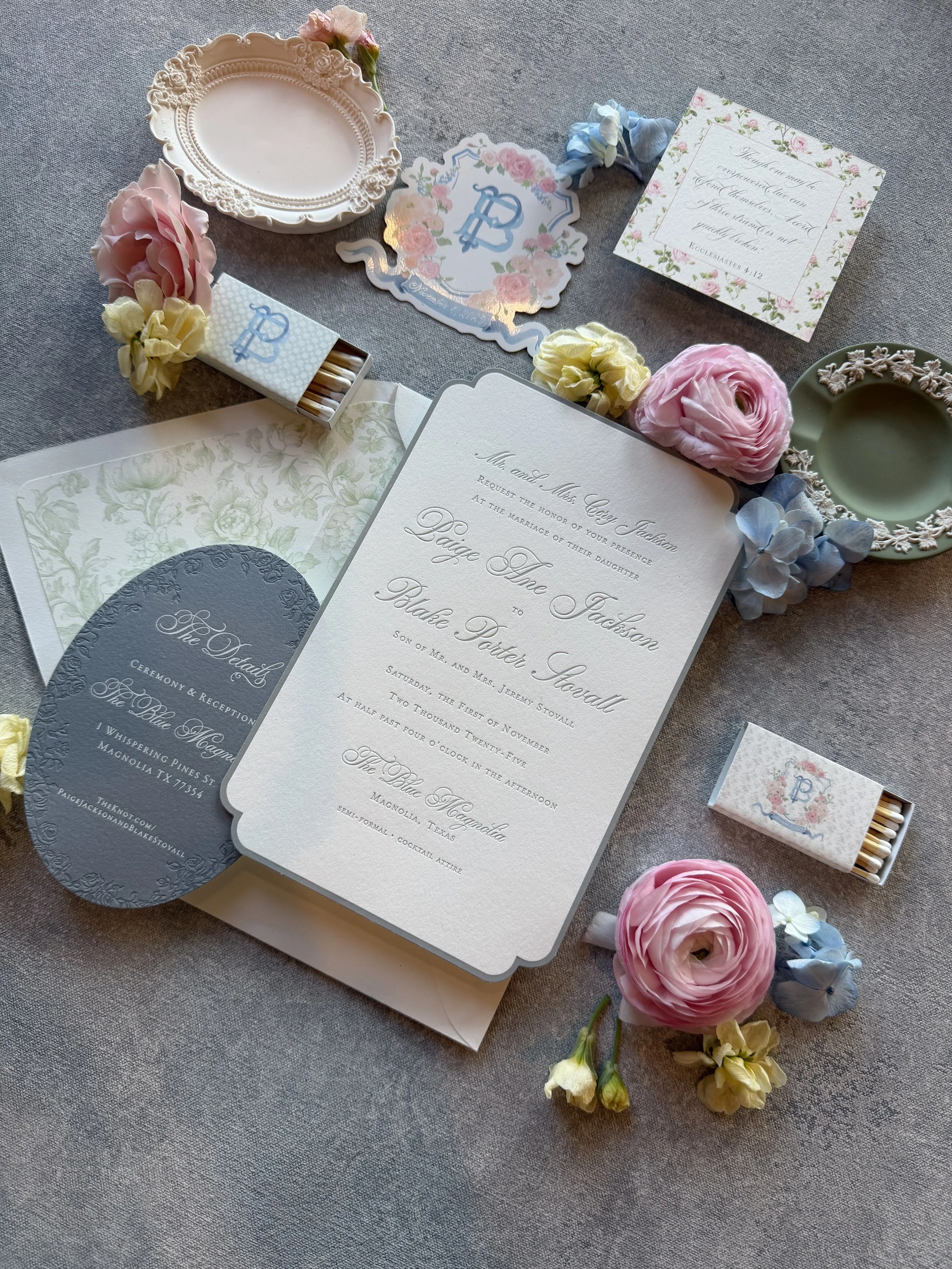 Wedding invitation, floral stationery, pastel-colored flowers, and matchboxes displayed on a table.
