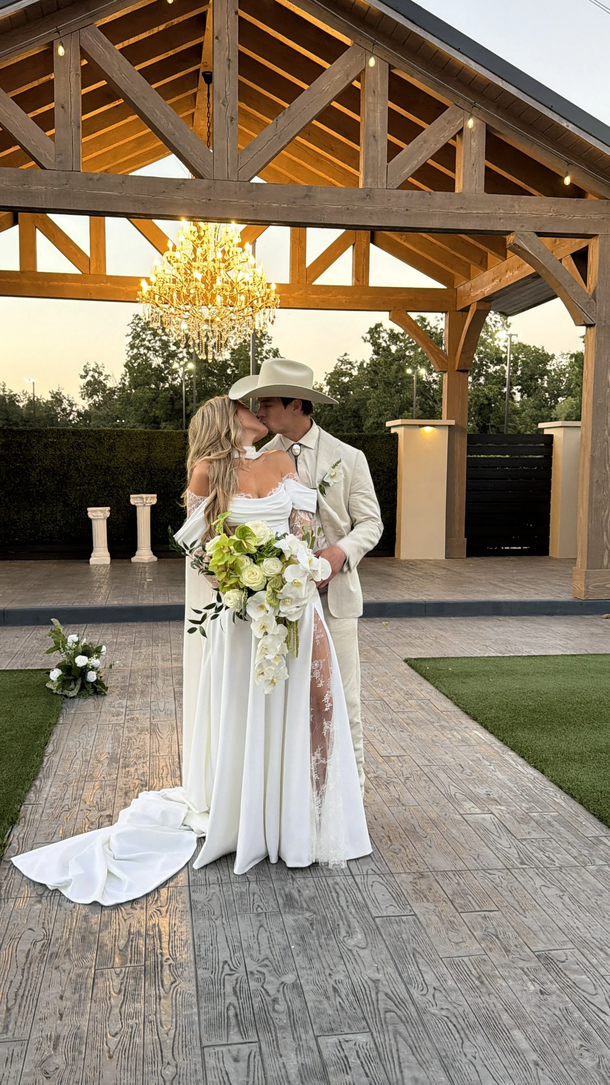 A bride and groom sharing a kiss at their wedding ceremony outdoors, with a wooden pavilion, chandelier, and greenery in the background.