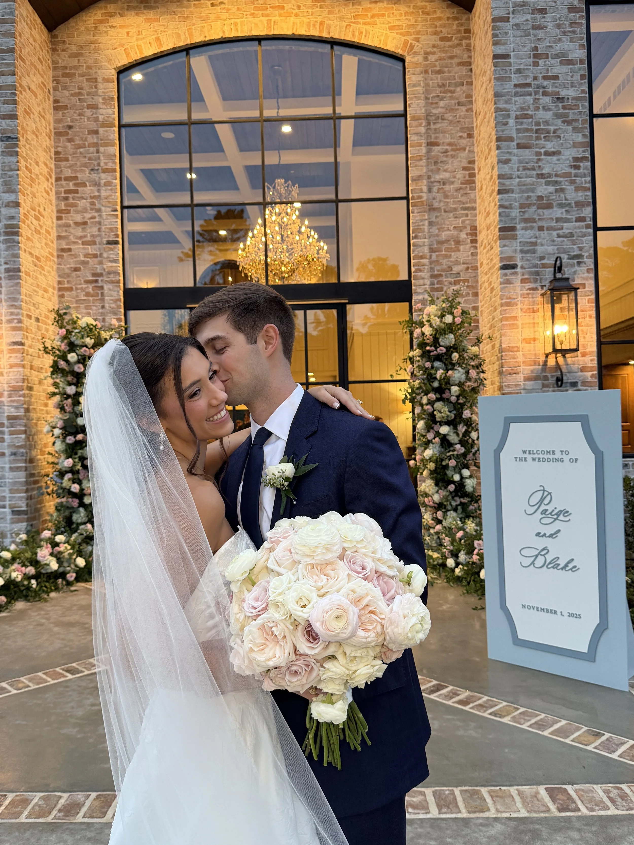 Bride and groom hugging, bride holding a bouquet of white and pink roses, standing in front of a decorated wedding arch with flowers, a large window with a chandelier inside, and a wedding sign with their names and wedding date.