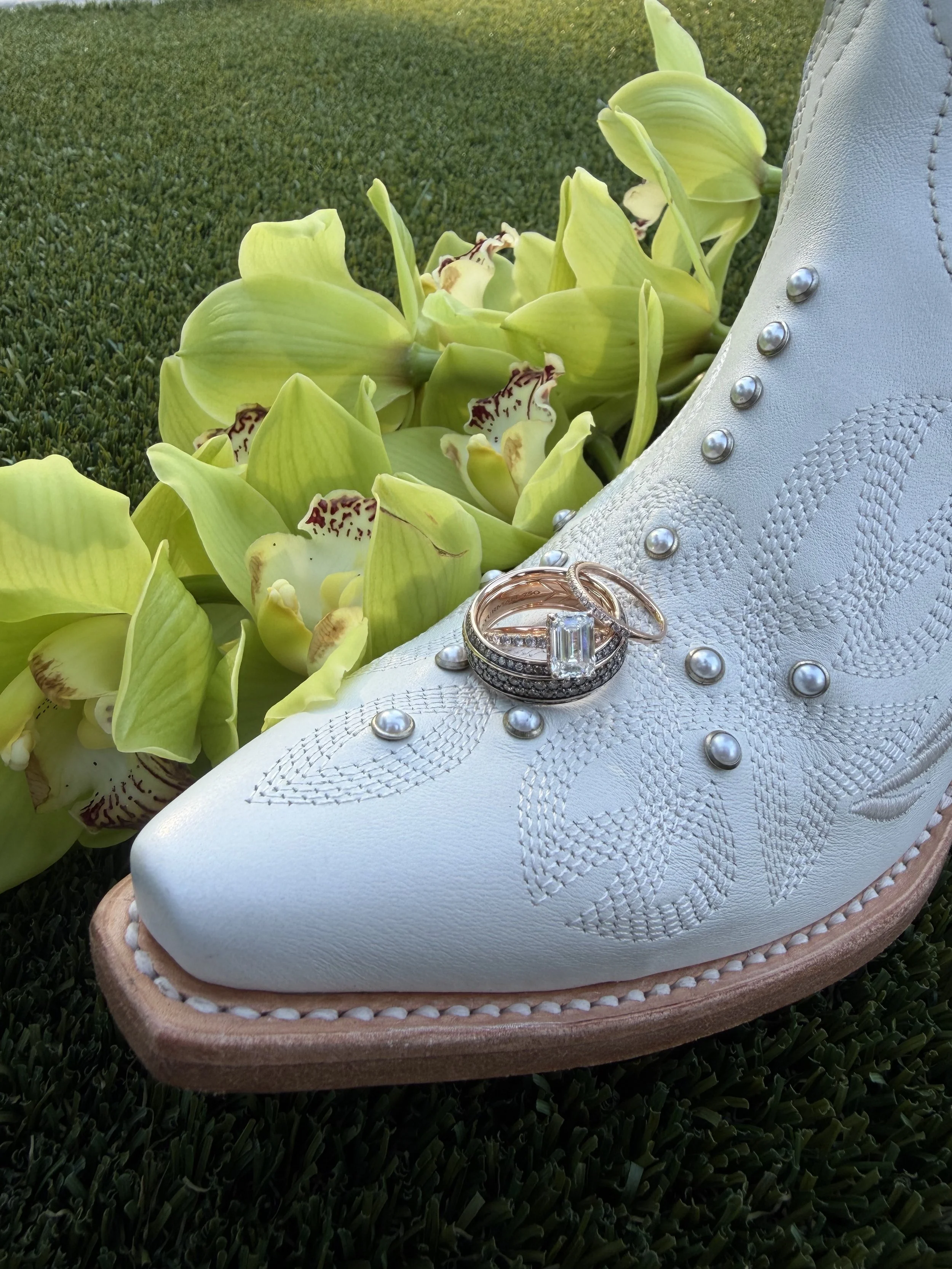 A white embroidered cowboy boot with pearl accents, adorned with four rings on top, surrounded by green orchids on a grassy surface.
