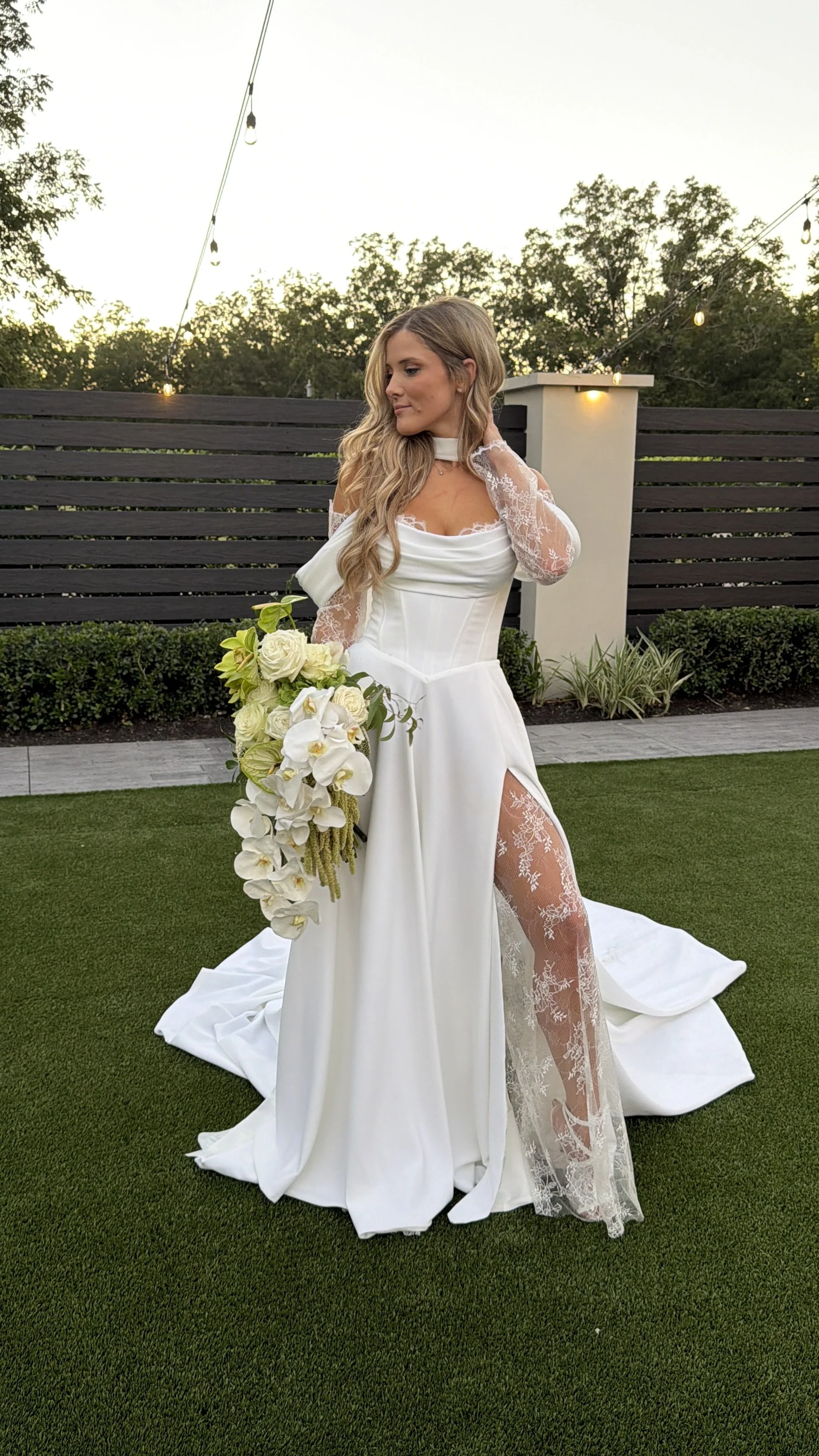 A bride in a white wedding dress with lace sleeves and a high slit, holding a cascading white and green floral bouquet, standing on a lawn during sunset, with a wooden fence, string lights, and trees in the background.