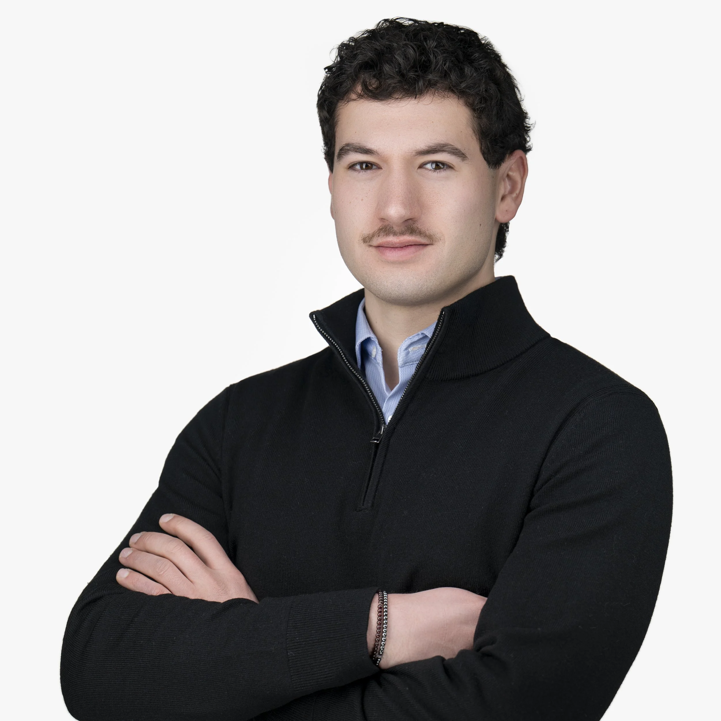 Portrait of a young man with dark curly hair, wearing a black zip-up sweater and light blue collared shirt, crossing arms, against a plain white background.