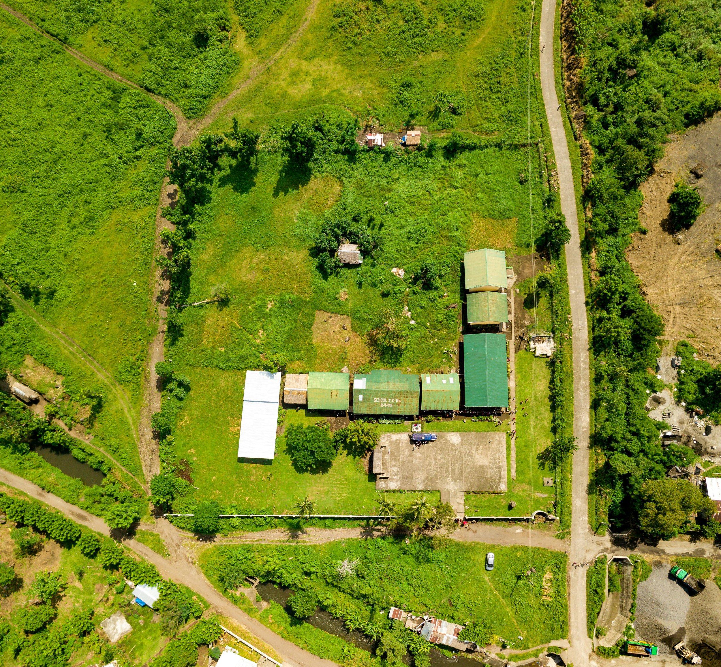 Aerial view of a rural area with green fields, scattered small buildings with green roofs, a forested area, and a dirt road running vertically on the right side.