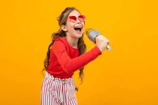 Young girl singing into a microphone against a yellow background, wearing sunglasses, a red top, and striped pants.