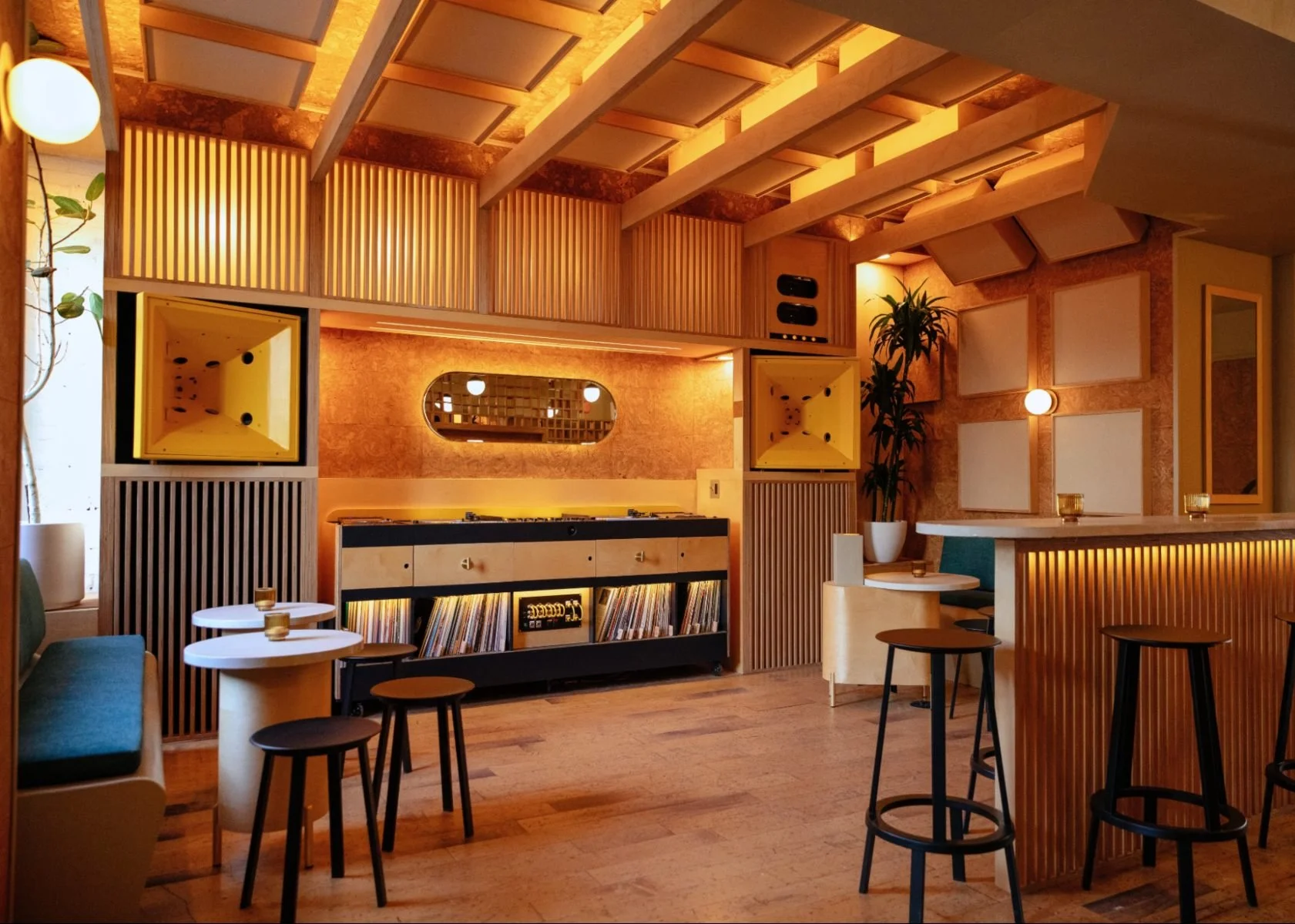 A cozy, modern cafe with warm lighting, wooden decor, small tables, bar stools, and a record player with vinyl records.