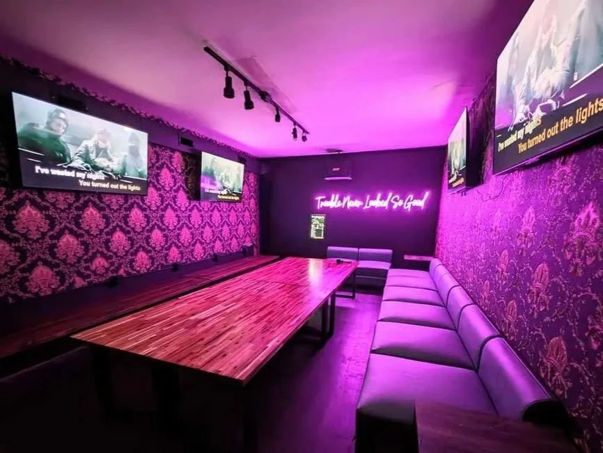 Empty karaoke room with purple neon lighting, patterned purple walls, multiple TVs with karaoke lyrics, and a long table with chairs along one side.
