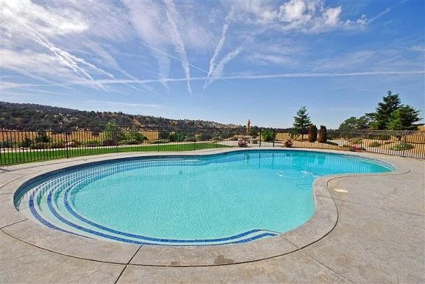 Empty outdoor swimming pool with blue water and a fence, landscape with hills and trees in the background, blue sky with clouds and contrails.