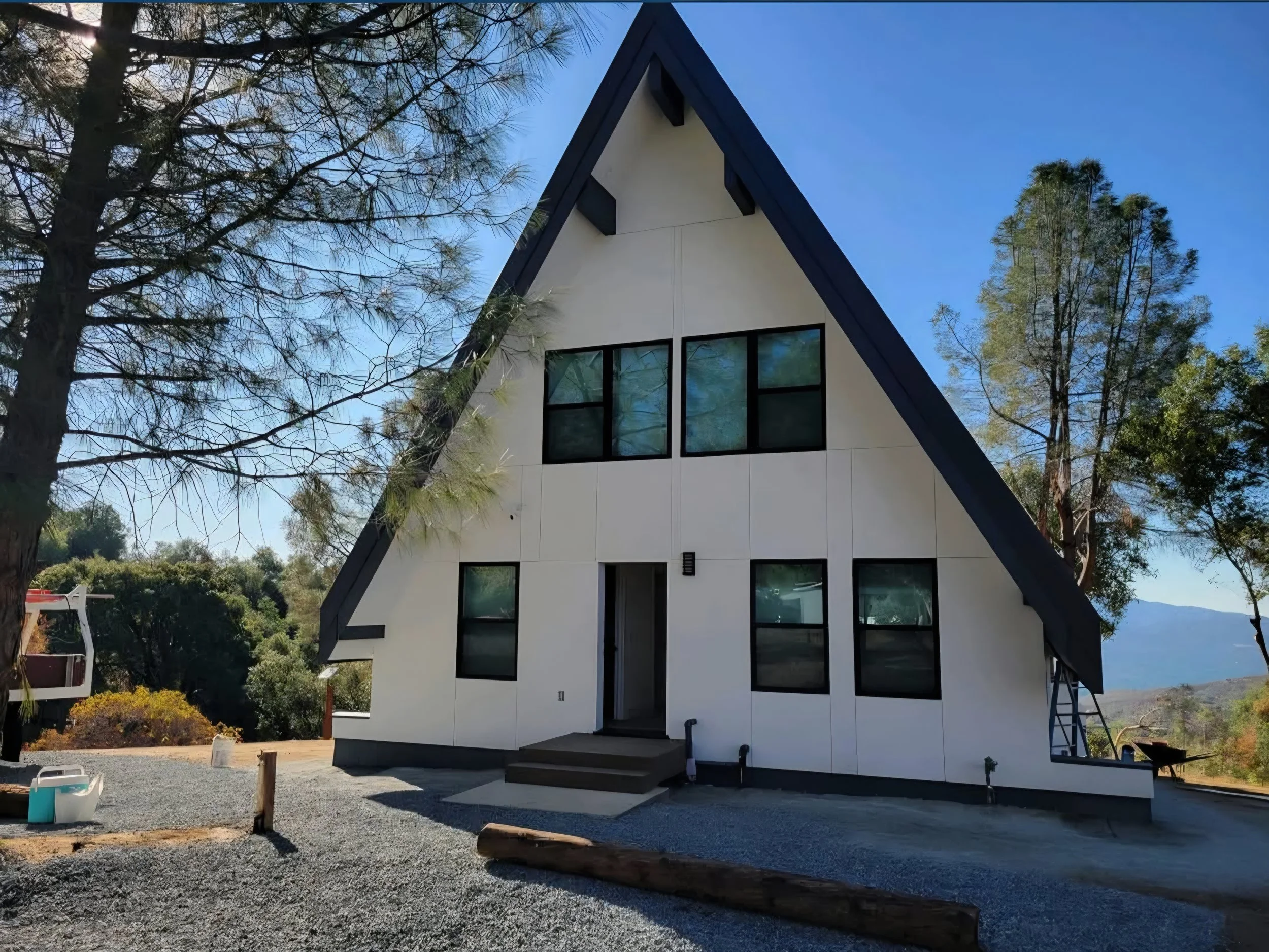 A modern A-frame house with white and black exterior, surrounded by trees and a gravel driveway, under a blue sky.