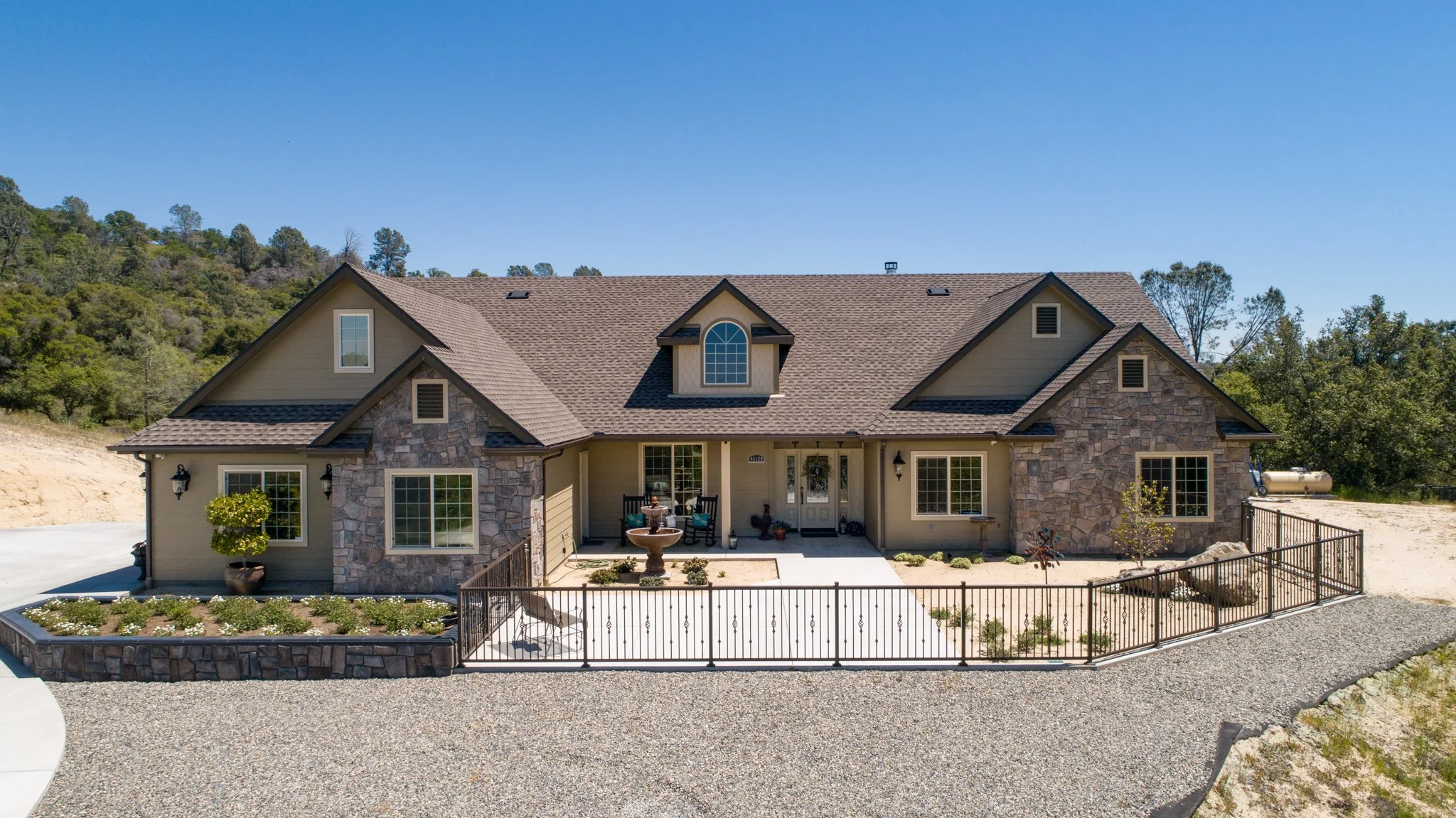 Large modern house with stone and beige siding exterior, multiple gabled rooflines, front porch with benches, decorative fountain, landscaped front yard with small trees and plants, surrounded by a gravel driveway, set against a background of green hills and a clear blue sky.