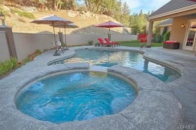 Backyard swimming pool with two umbrellas, lounge chairs, and a hot tub with bubbling water, surrounded by a concrete patio and a fence.
