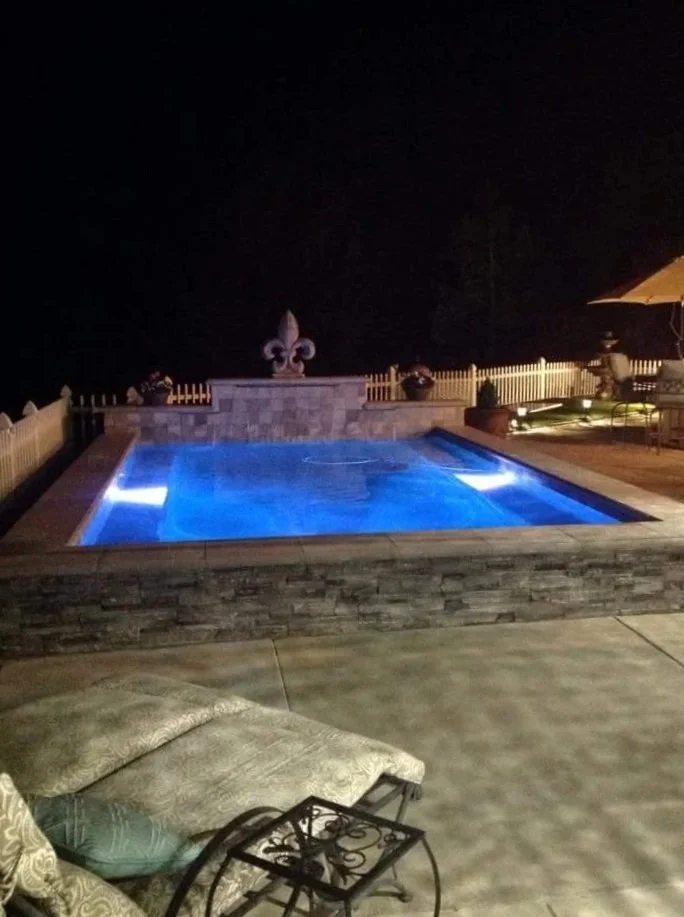Nighttime view of illuminated outdoor swimming pool surrounded by a patio with furniture and decorative fencing.