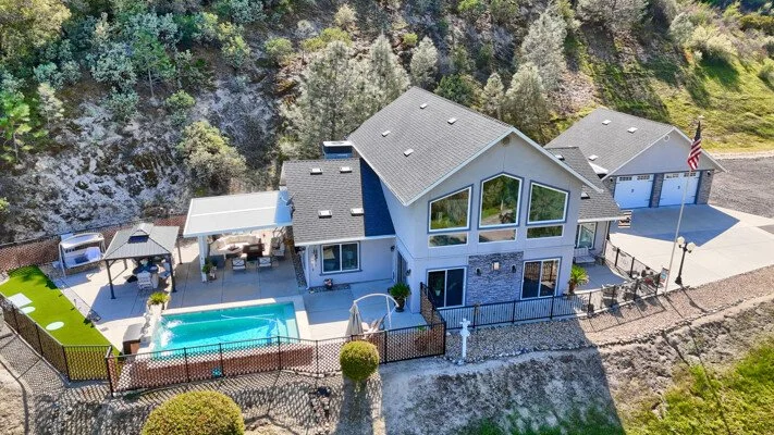 A modern two-story house with a backyard pool, patio area with outdoor furniture, and a separate garage, surrounded by a fenced yard and located near a hillside with trees.