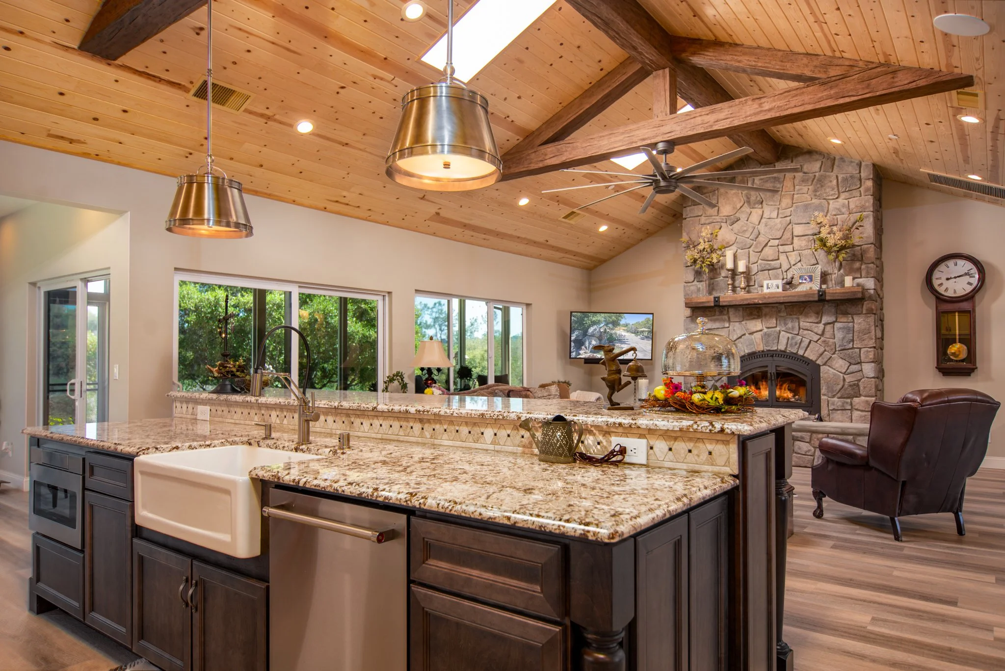 Open kitchen and living room with wood vaulted ceiling, stone fireplace, granite countertops, island with sink, leather armchair, wall clock, and large windows showing outdoor greenery.