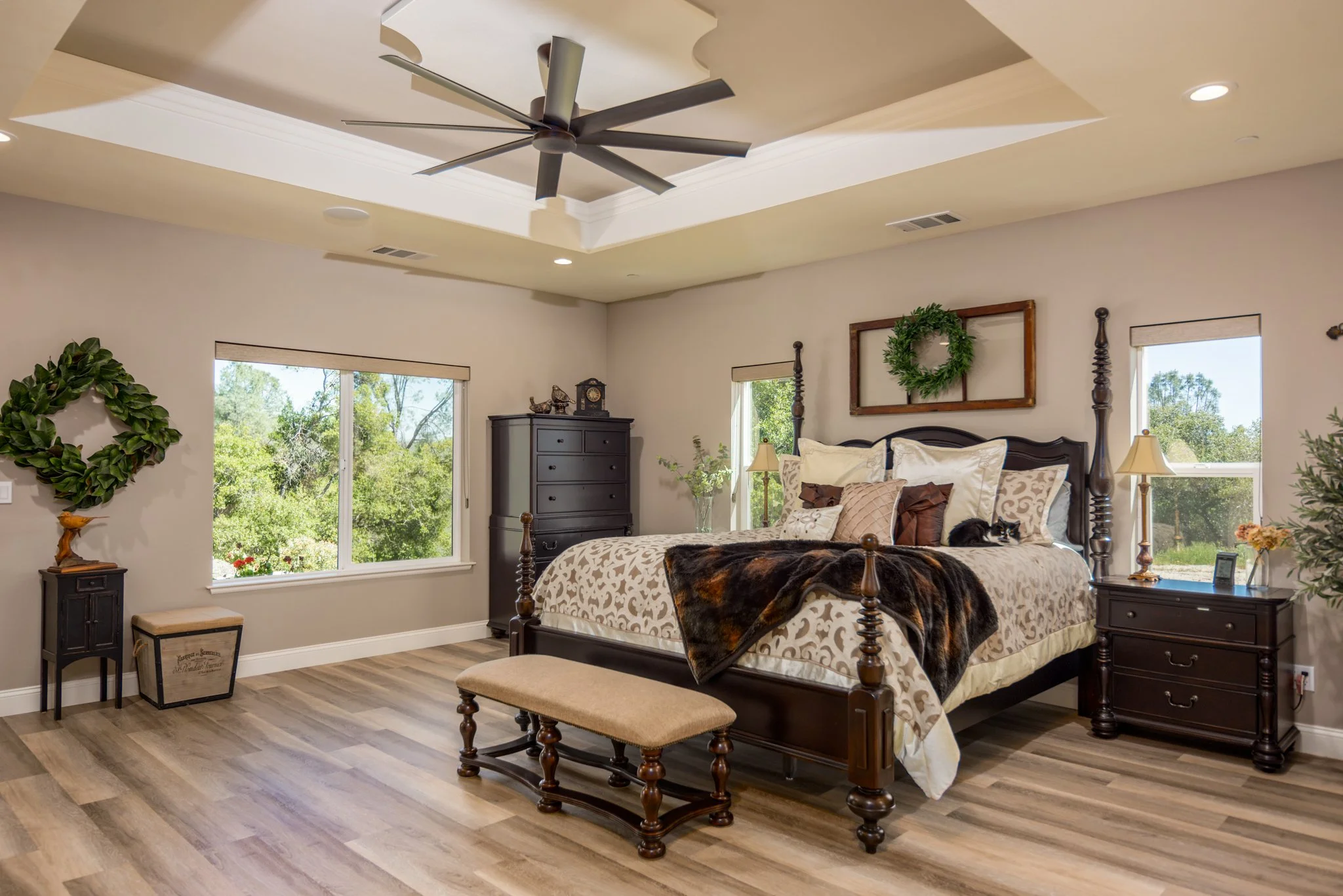 A spacious bedroom with a large four-poster bed, decorated with multiple pillows and a fur throw, a bench at the foot of the bed, and nightstands on either side with lamps. There are windows revealing green trees outside, and the room features a tray ceiling with a ceiling fan, hardwood floors, and wall decor including a wreath and framed art.