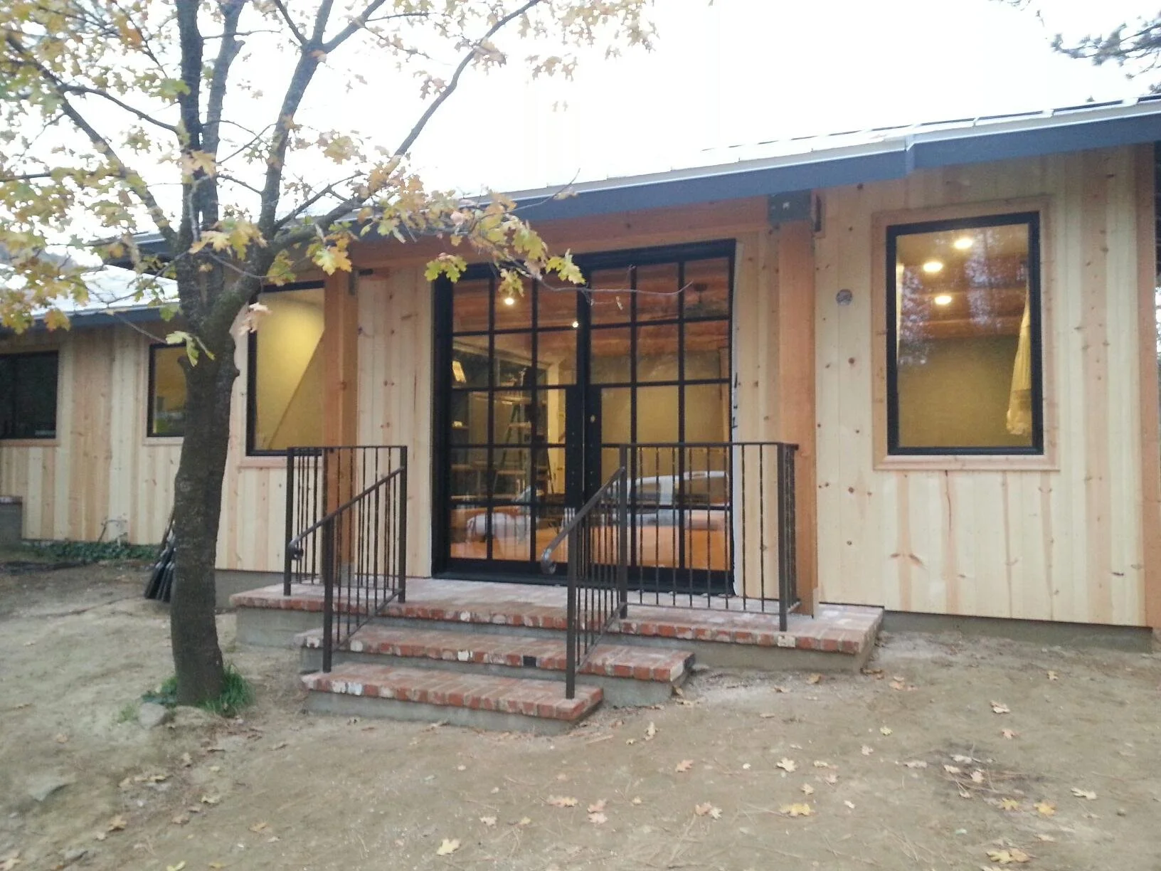 front of a newly constructed house with wooden exterior, black metal staircase and railing, brick steps, large glass door, and windows, surrounded by a dirt yard and trees.