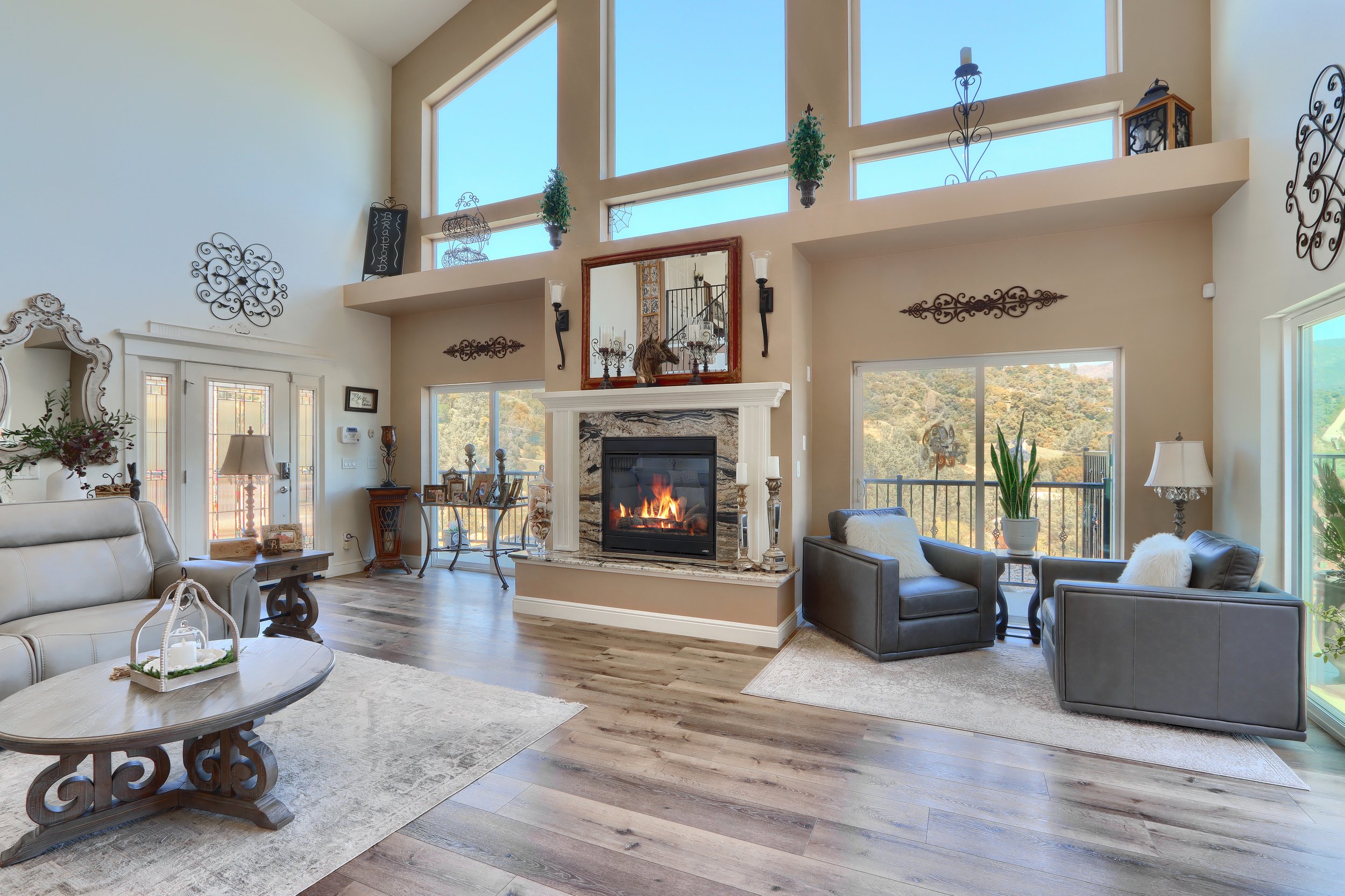 A spacious living room with high vaulted ceilings and large windows showing a scenic view of hills. The room features a fireplace with a mirror above it, surrounded by armchairs, sofas, and various decorative items, creating a cozy and elegant atmosphere.