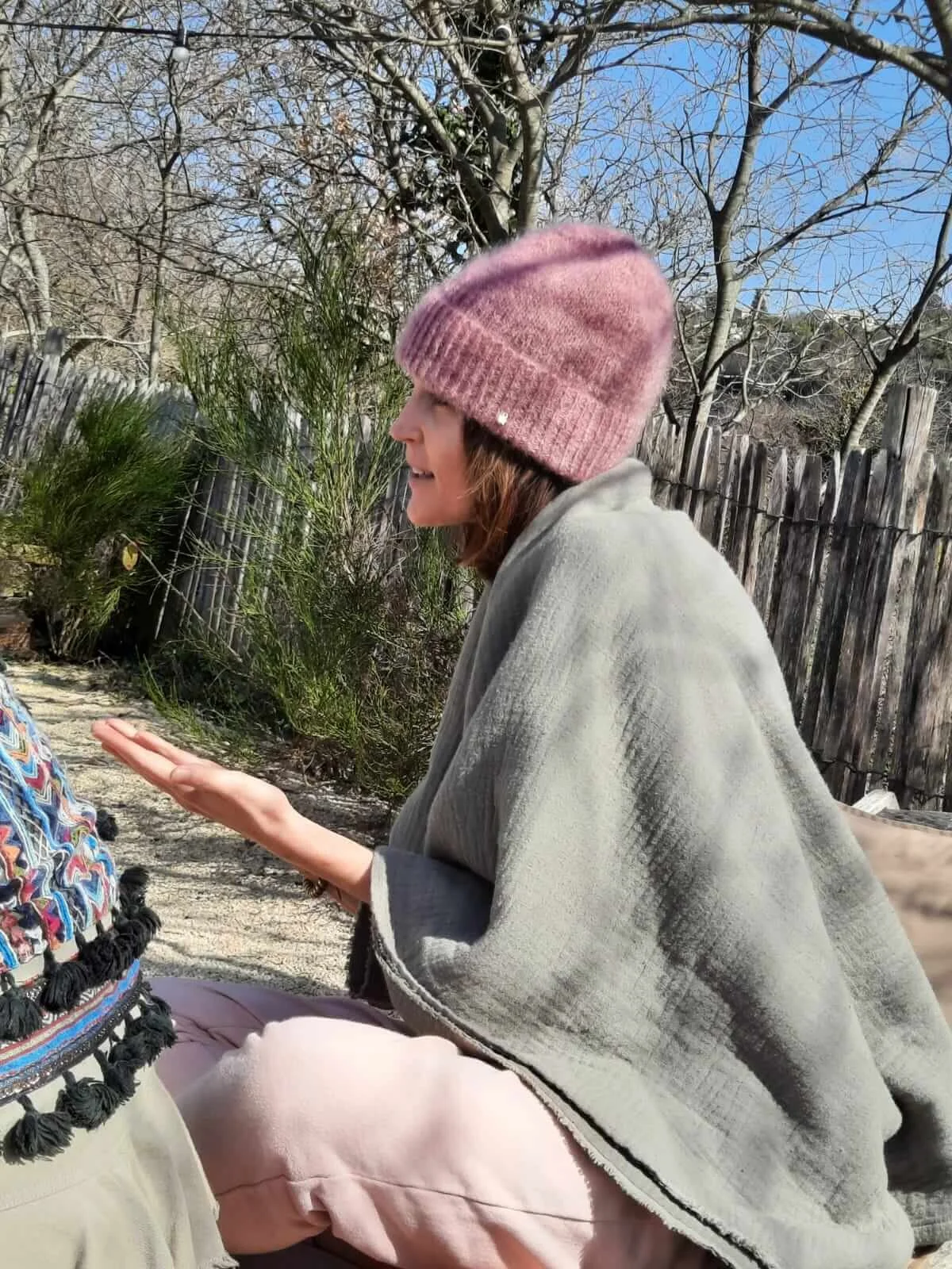 Ludivine, souriante portant un bonnet rose en laine, un poncho gris clair, assise dans un jardin avec un arbre sans feuilles et une clôture en bois en arrière-plan.