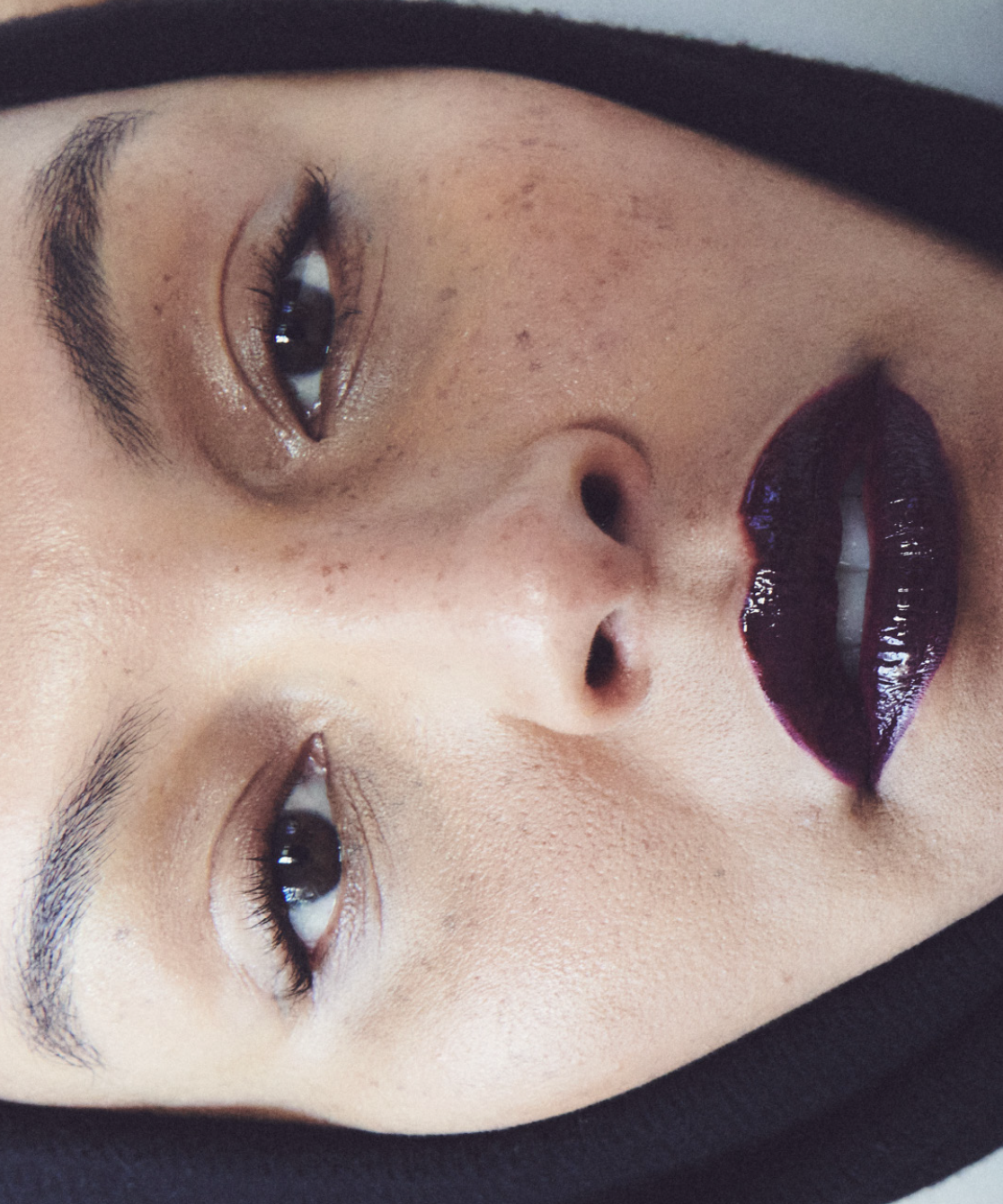 Close-up of a person's face with dark lipstick, natural eye makeup, and a dark head covering.