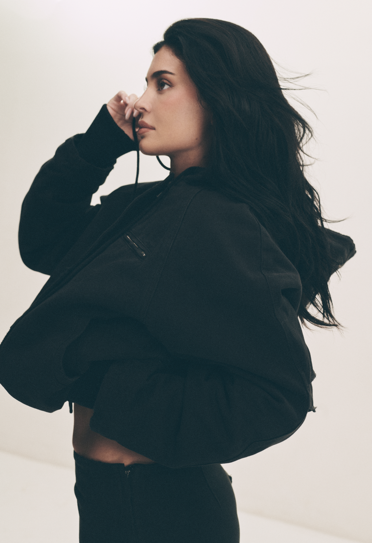 A woman with long dark hair wearing a black jacket and black pants, sitting against a plain light background, holding a headphone cord near her face.