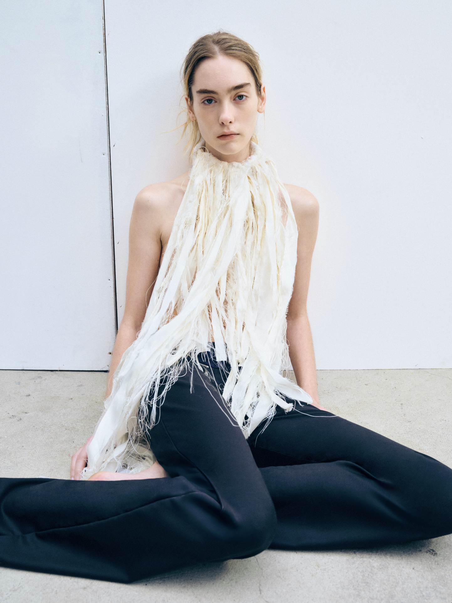 A woman with blonde hair sitting on the ground in front of a white wall, wearing a feathered, white top and black pants.