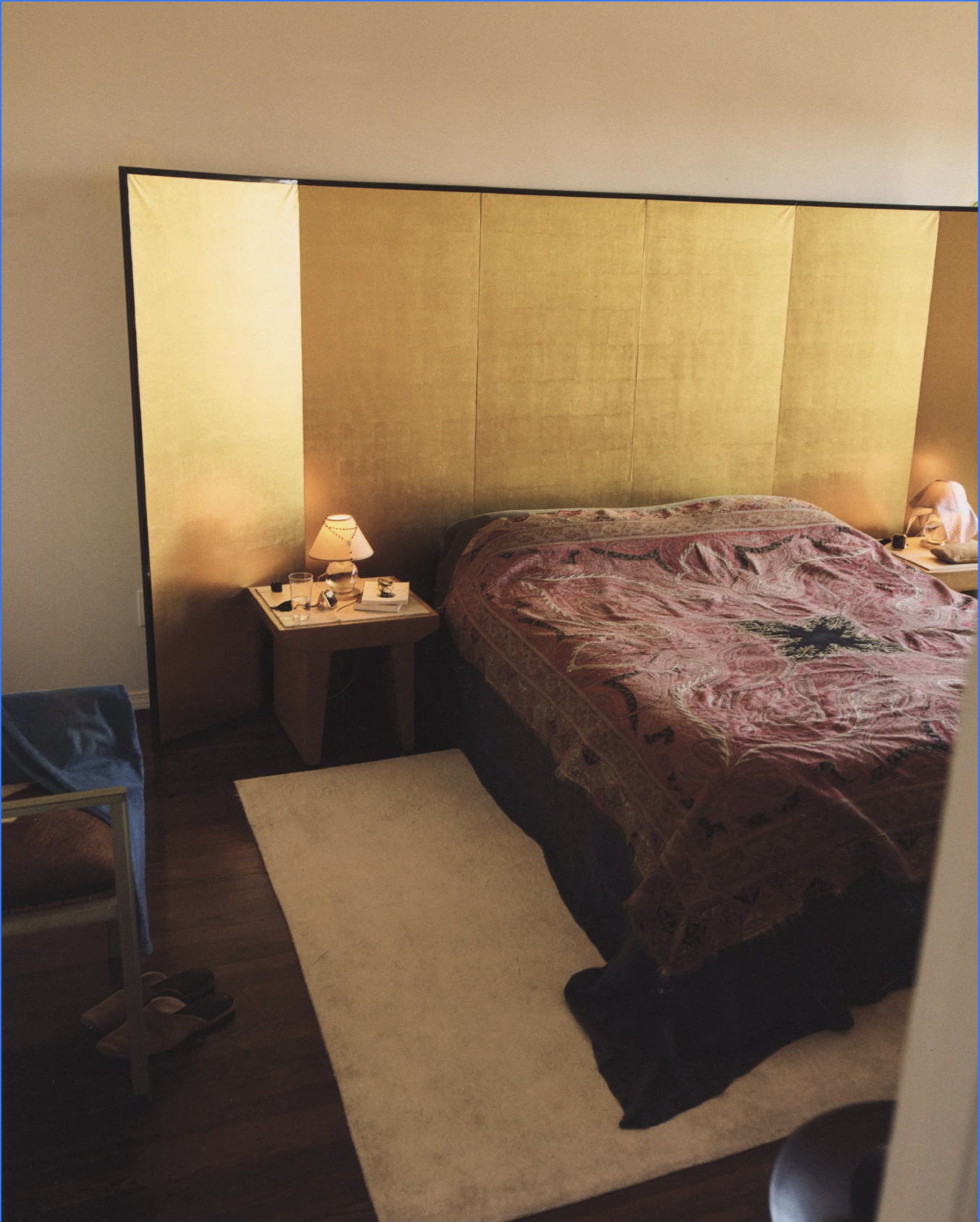 A bedroom with a large bed covered by a patterned bedspread, two bedside tables with lamps, a small chair, and a beige rug on dark wooden floor. A gold-colored headboard extends across the wall behind the bed.