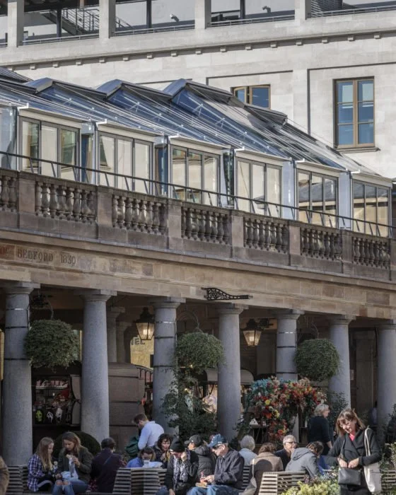 People sitting at an outdoor cafe with columns, flowers, and a glass greenhouse structure above, located next to a stone building.