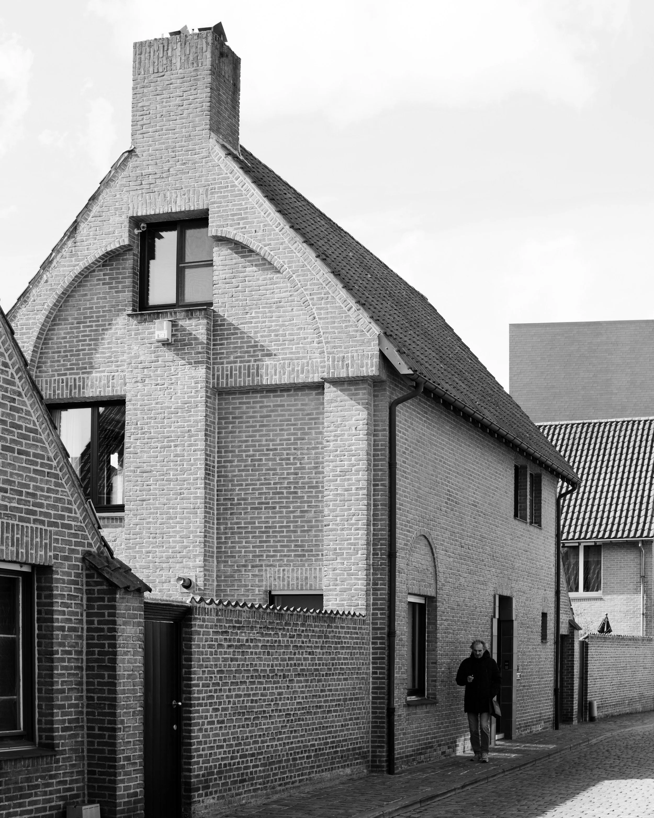 A black and white photo of a brick house with a sloped roof and chimney, with a person walking on the sidewalk in front.