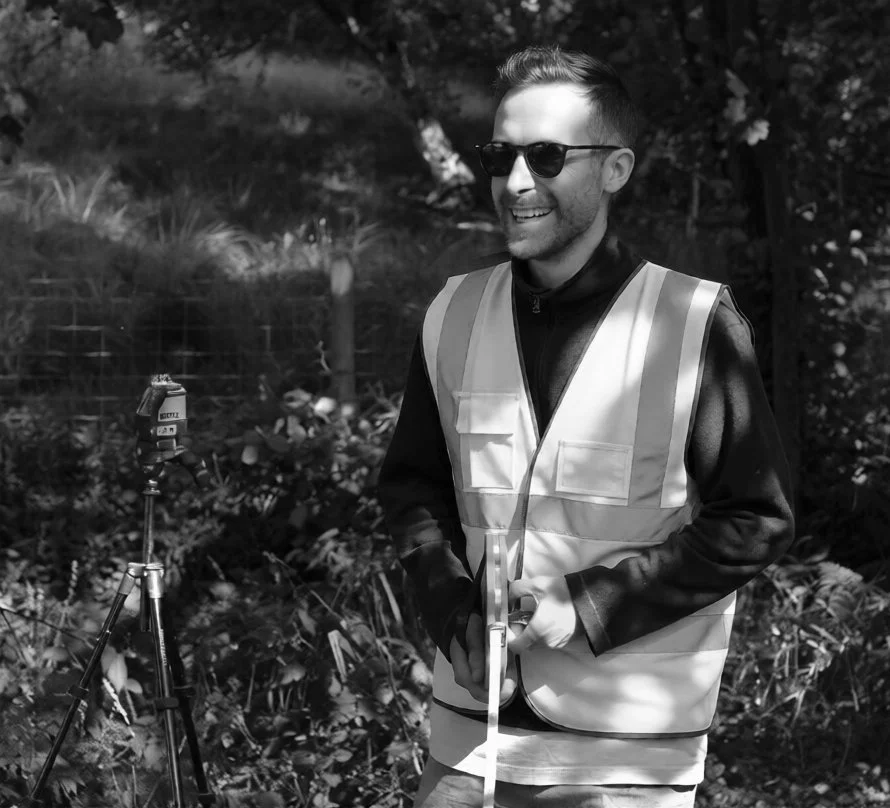 A smiling man wearing sunglasses and a reflective safety vest standing outdoors with camera equipment on a tripod, surrounded by trees and foliage.
