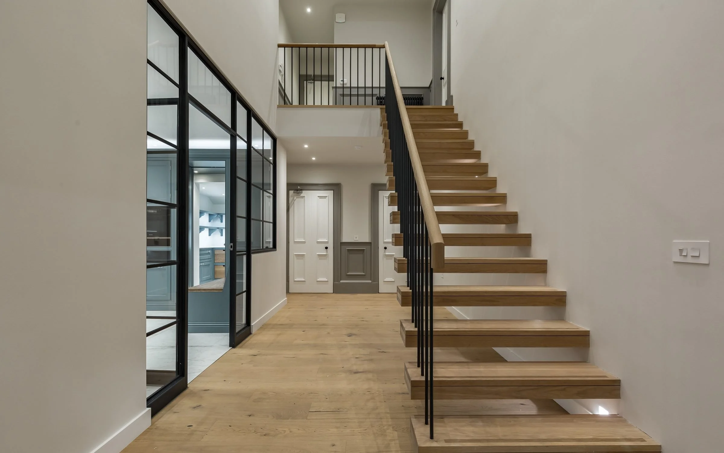 Modern interior with wooden staircase, black metal railing, glass partition, following with a hallway and doors, light-colored walls, subway tile floor visible in adjoining room.