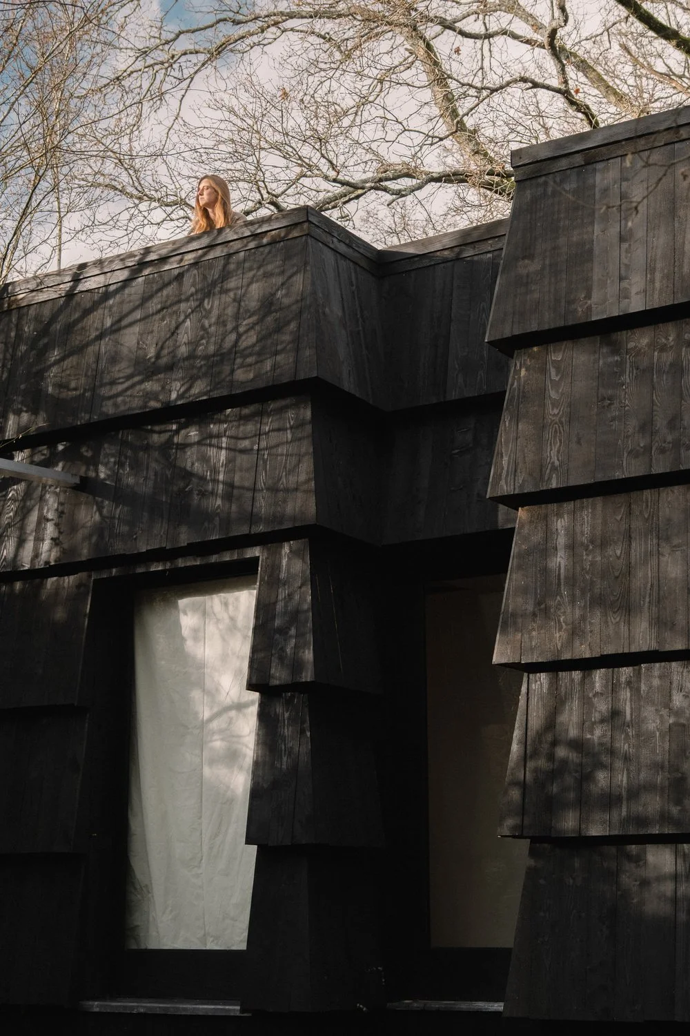 A young woman with long hair looking out from an elevated black wooden balcony of a modern building with an angular design, surrounded by leafless trees and a clear sky.