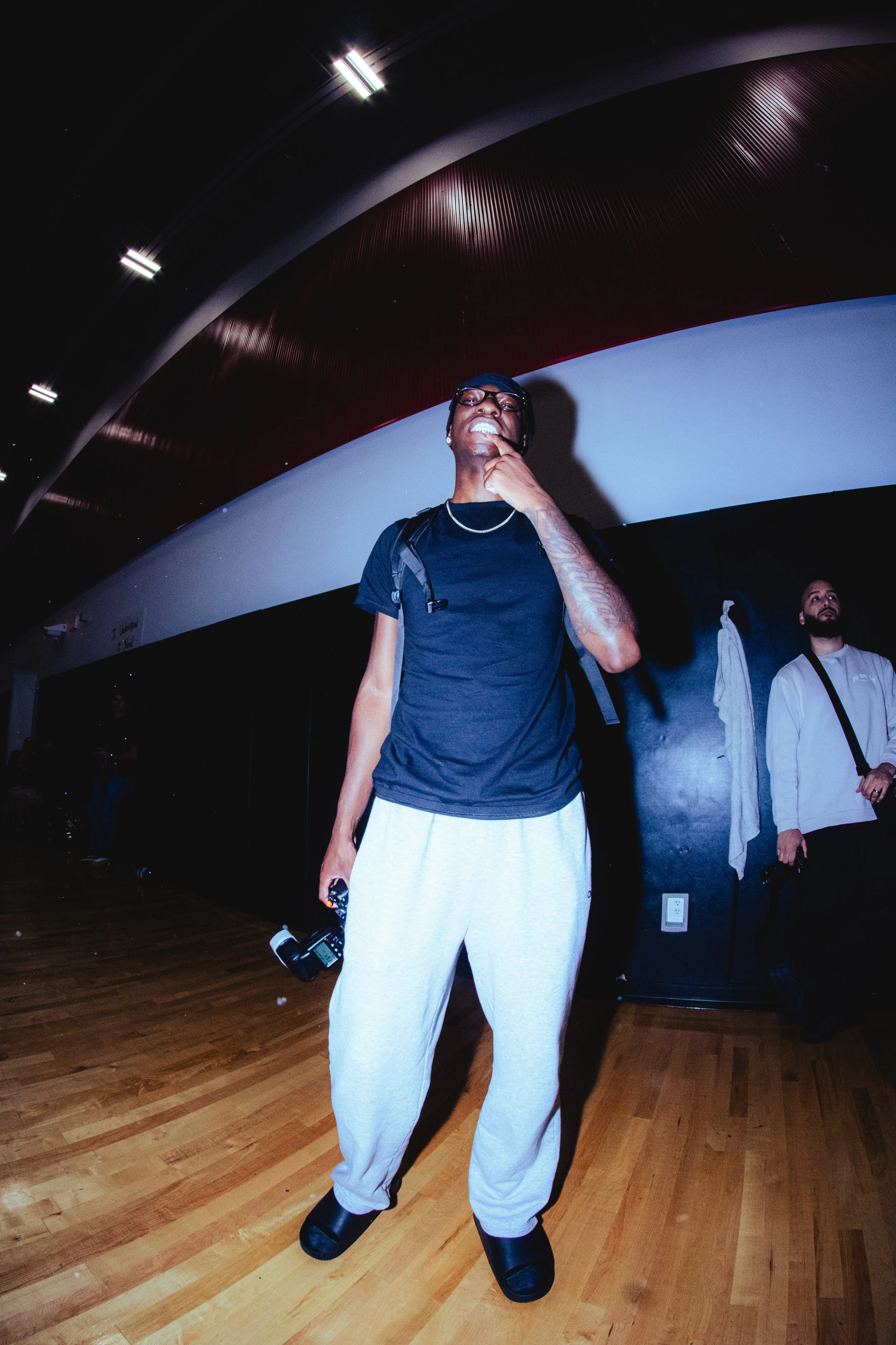 A man in black t-shirt, gray sweatpants, and slides standing in a room with wooden floors and curved ceiling, holding a camera, with another man in white standing behind him.