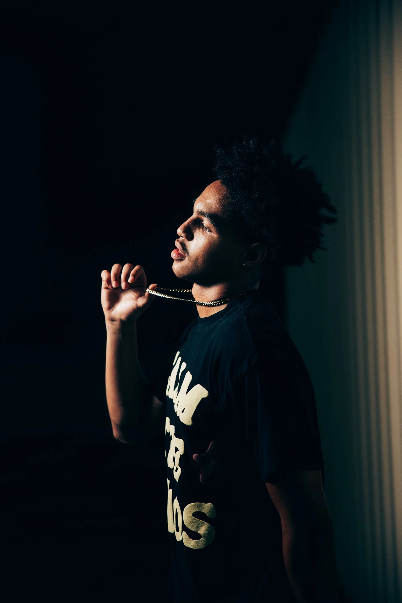 A young man with styled hair standing in profile against a dark background, holding a gold chain necklace near his neck, wearing a black T-shirt with partially visible white text.