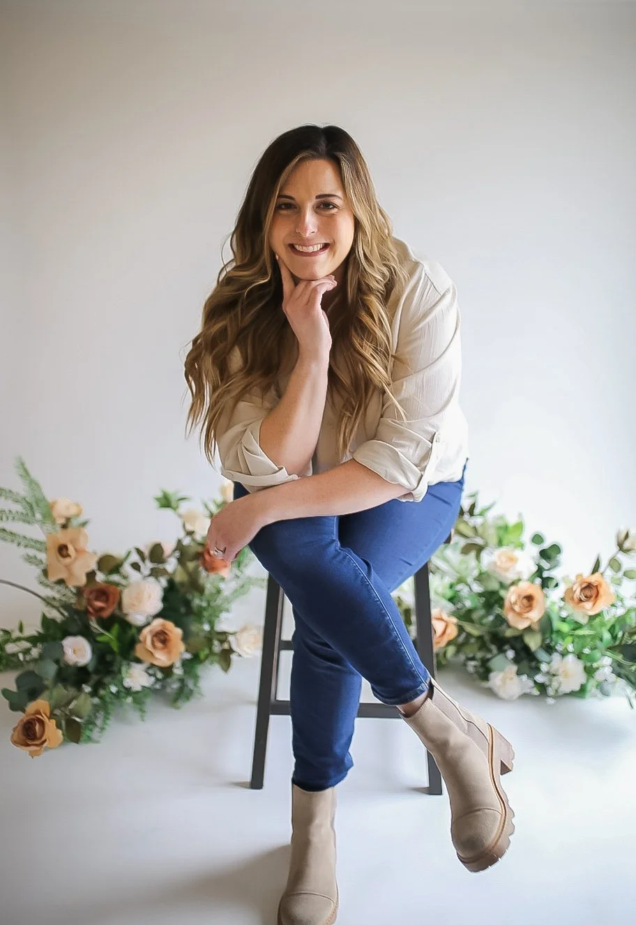 A woman with long wavy hair, wearing a cream blouse and blue jeans, sits on a stool surrounded by peach, cream, and white flowers, smiling at the camera.