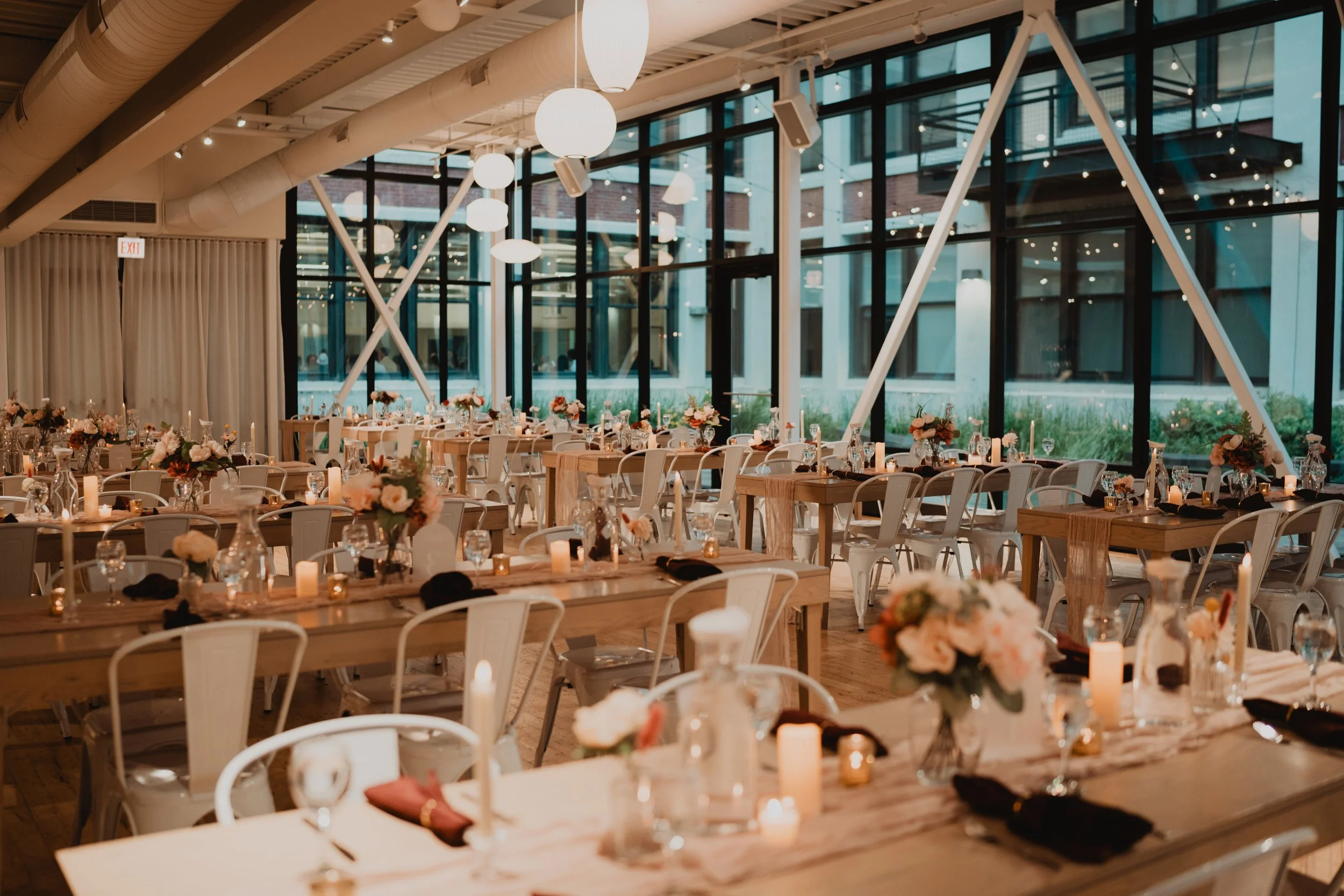 Elegant event space with wooden tables decorated with floral centerpieces, candles, and glassware, surrounded by white chairs, with large windows and string lights creating a warm atmosphere.