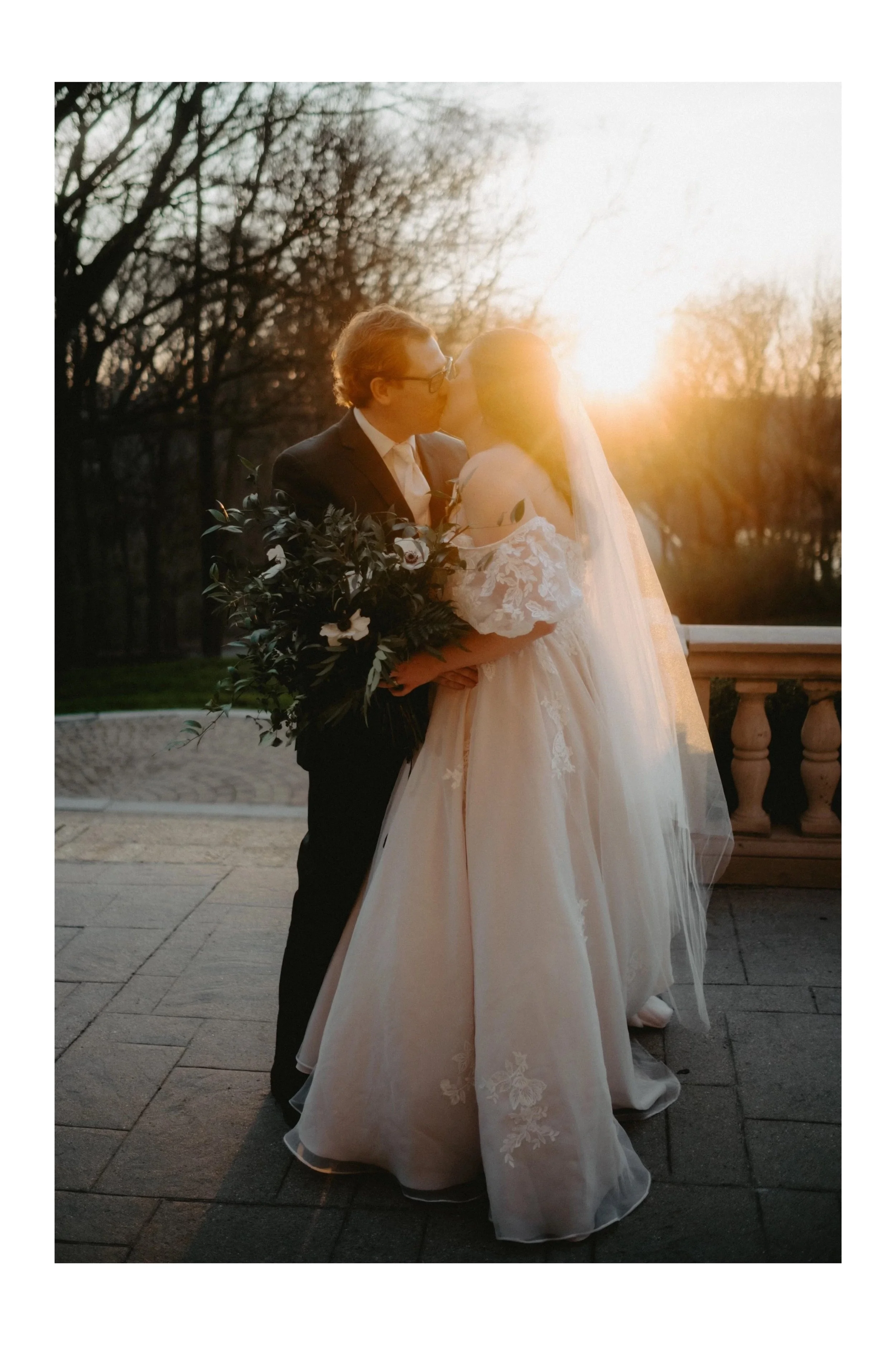 A newlywed couple sharing a kiss during sunset on a terrace, with trees in the background.