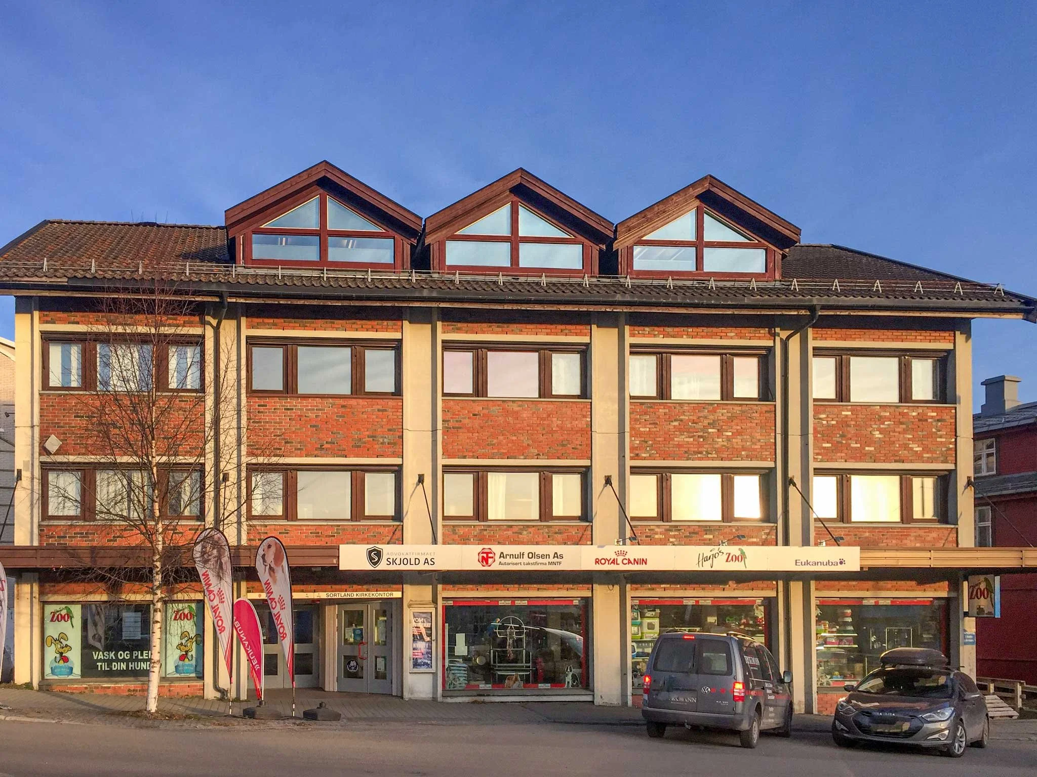 Fleretasjes bygård på Sortland med butikk i første etasje, butikkvindu med kaker foran og parkering foran bygningen. Kvartal 20 AS.