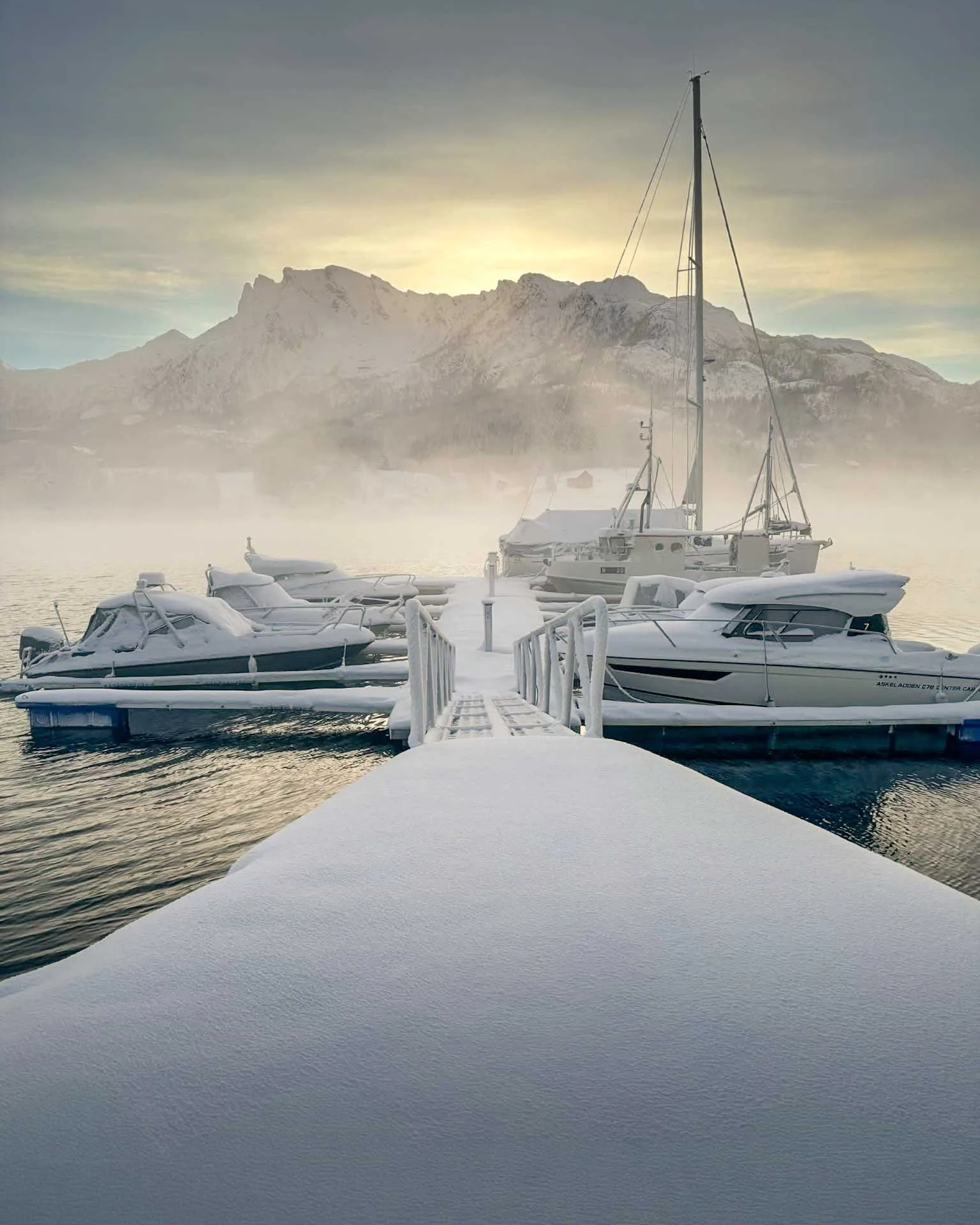 Snødekt brygge i Eidsfjorden, med båter foran fjell og tåke i bakgrunnen under en soloppgang eller solnedgang. Kvartal 20 AS.