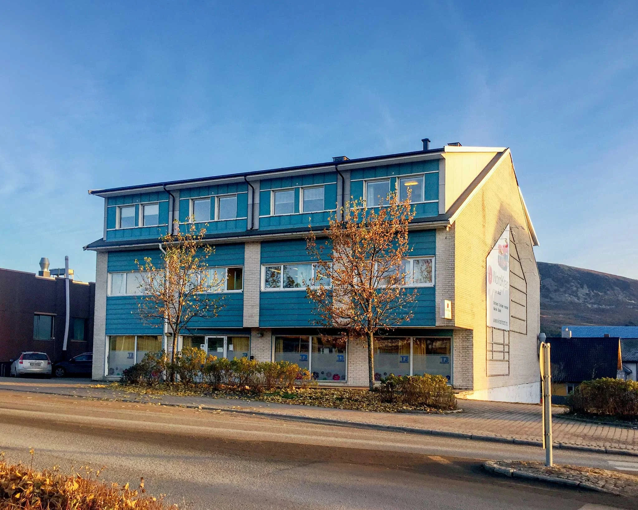 Tre etasjes bygning på Sortland, med blå og hvit fasade, to busker og biler parkert ved siden av. Skogen og fjell i bakgrunnen.. Kvartal 20 AS.