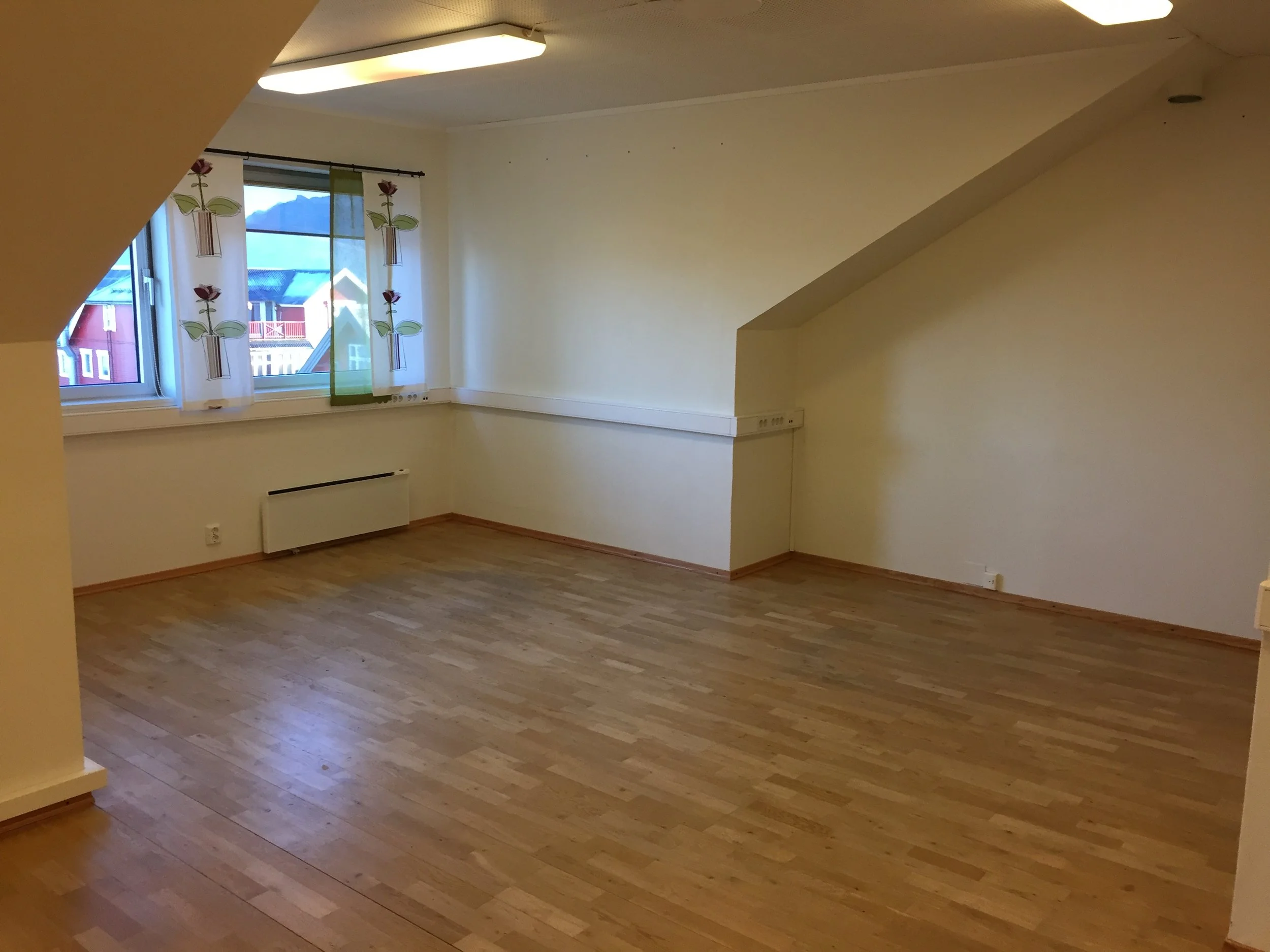 Et tomt kontor med tre vinduer, hvite vegger, tregulv, og en skjev takoverflate.. Kvartal 20 AS - Sortland.