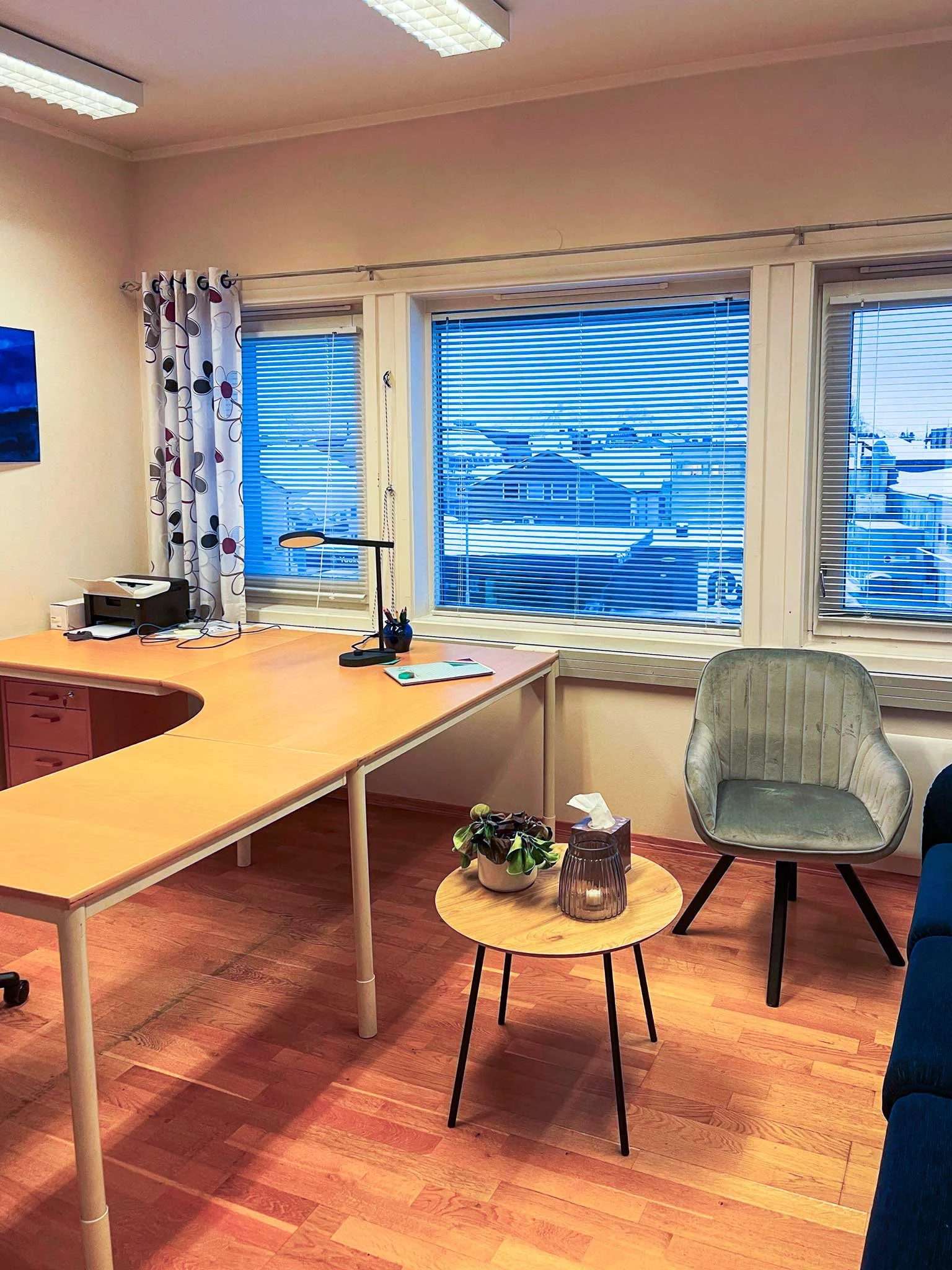 Et kontor på Sortland med et hjørnebord, en grøn lenestol, et lite rundt bord med en plante, en lys lampe, og et håndkle på et glasskrus. Foran vinduene er det vertikale blinds og en gardin med blomsterprint.. Kvartal 20 AS.