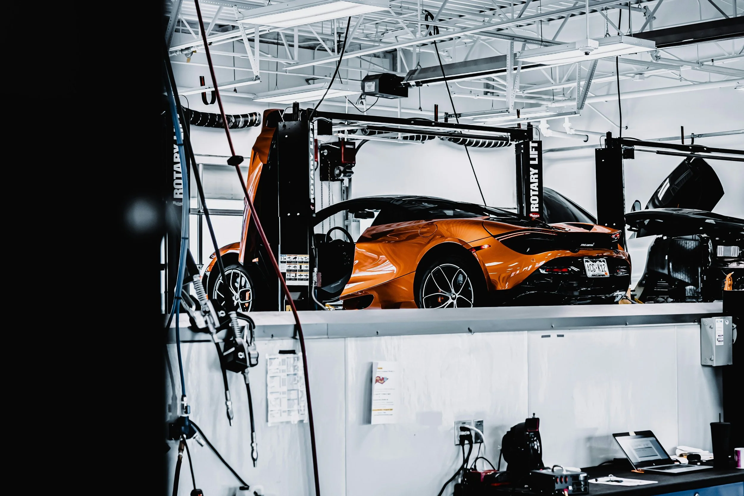 An image of a garage with a bright orange sports car on a lift, surrounded by equipment and tools.