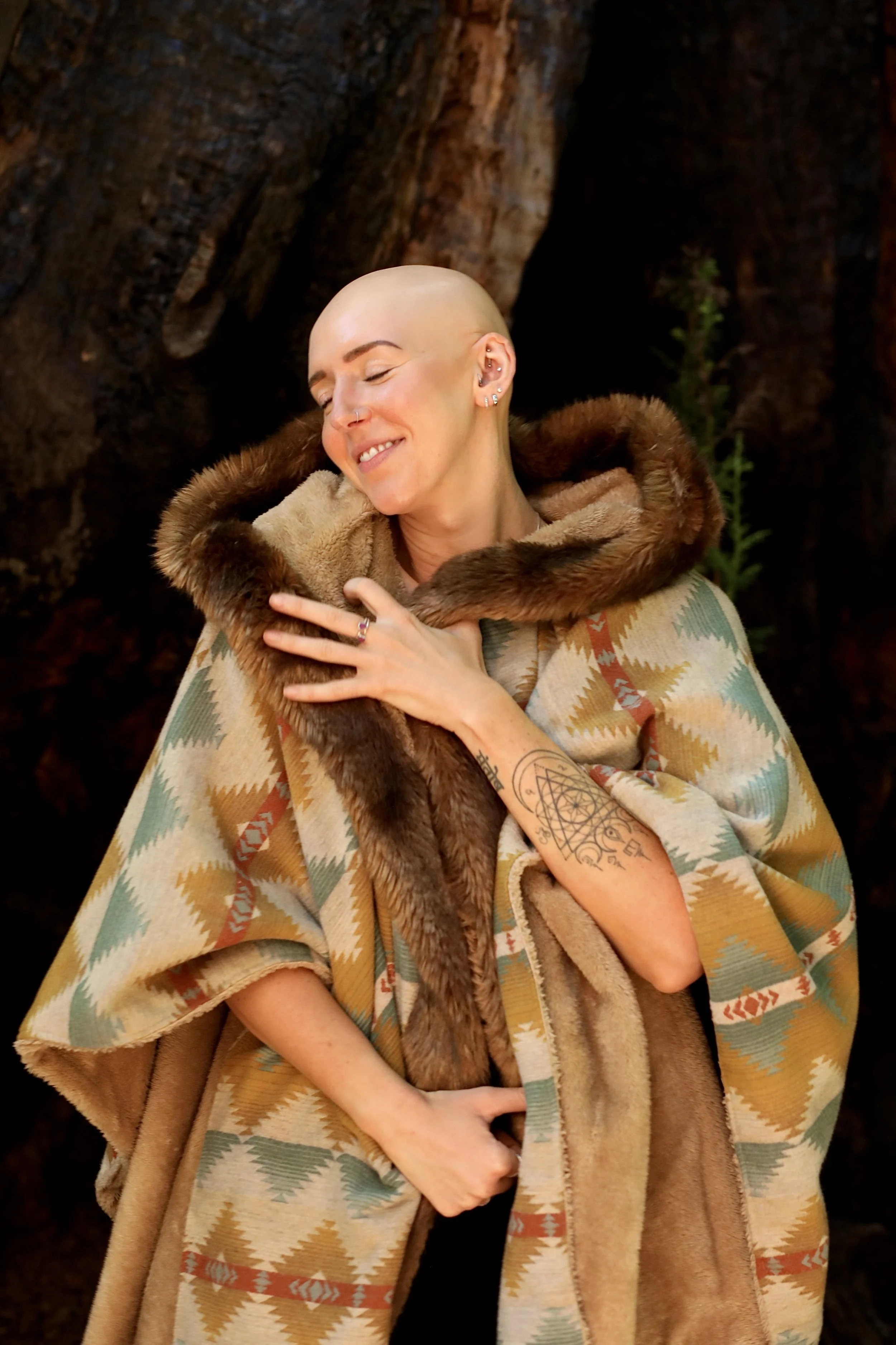 A woman with a shaved head, multiple earrings, and tattoos on her arm, smiling with eyes closed, wearing a patterned shawl with a fur-lined collar, standing in front of a large tree trunk with green plants in the background.