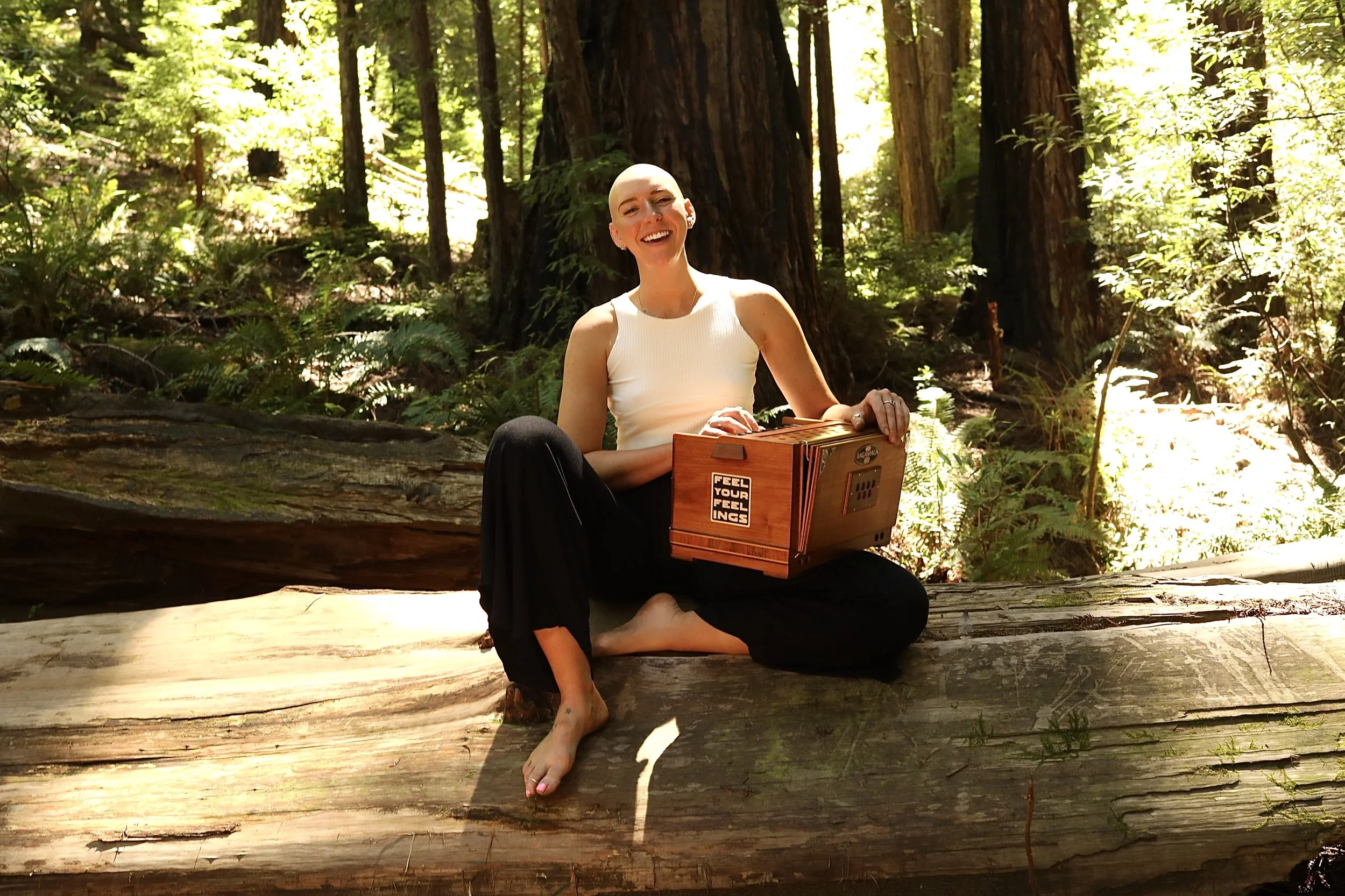 A woman with a shaved head smiling while sitting on a log in a forest, holding a wooden cajón musical instrument.